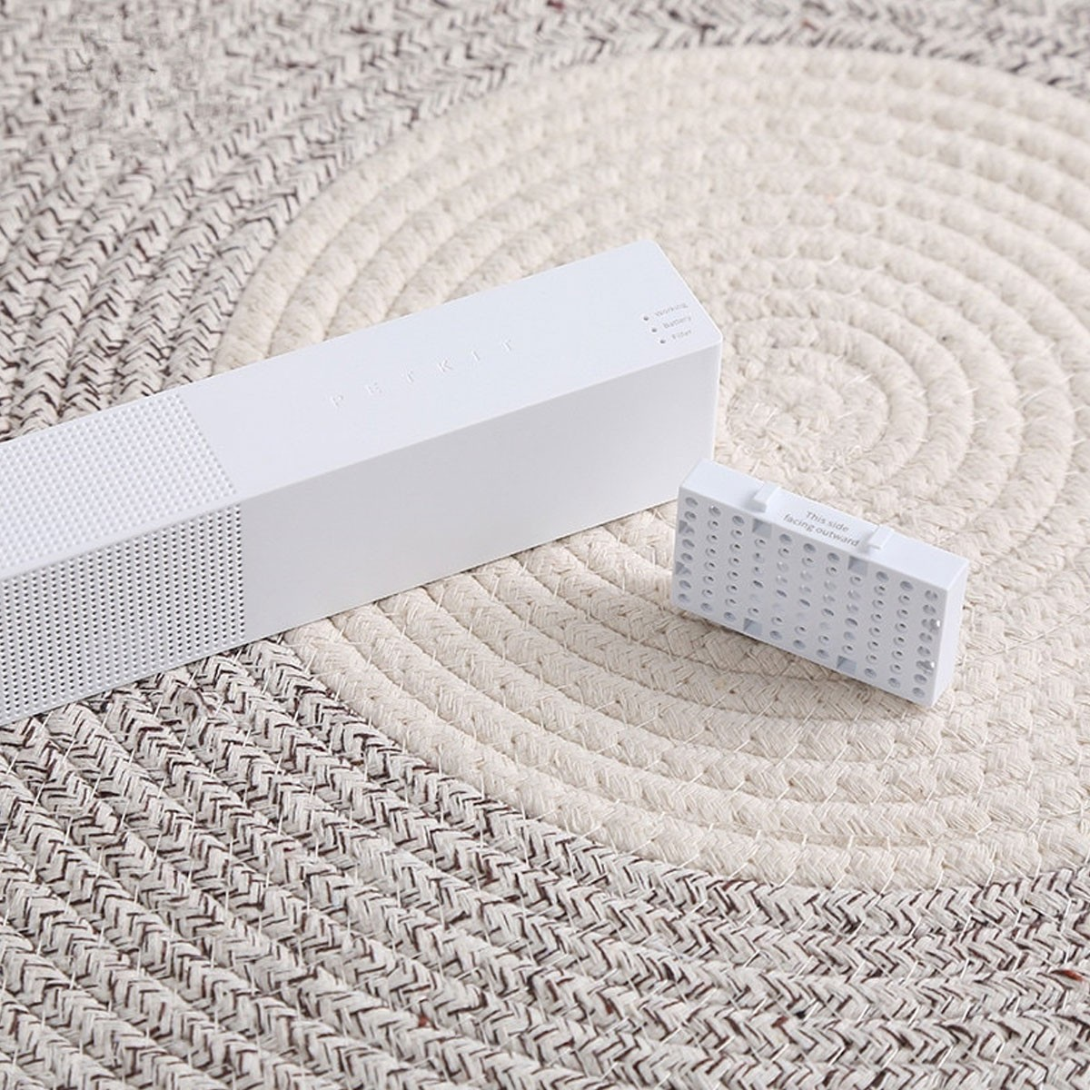 Białe urządzenie Petkit i mały prostokątny przedmiot na beżowo-brązowej tkanej macie. Na małym przedmiocie napis 'Ta strona na zewnątrz'.