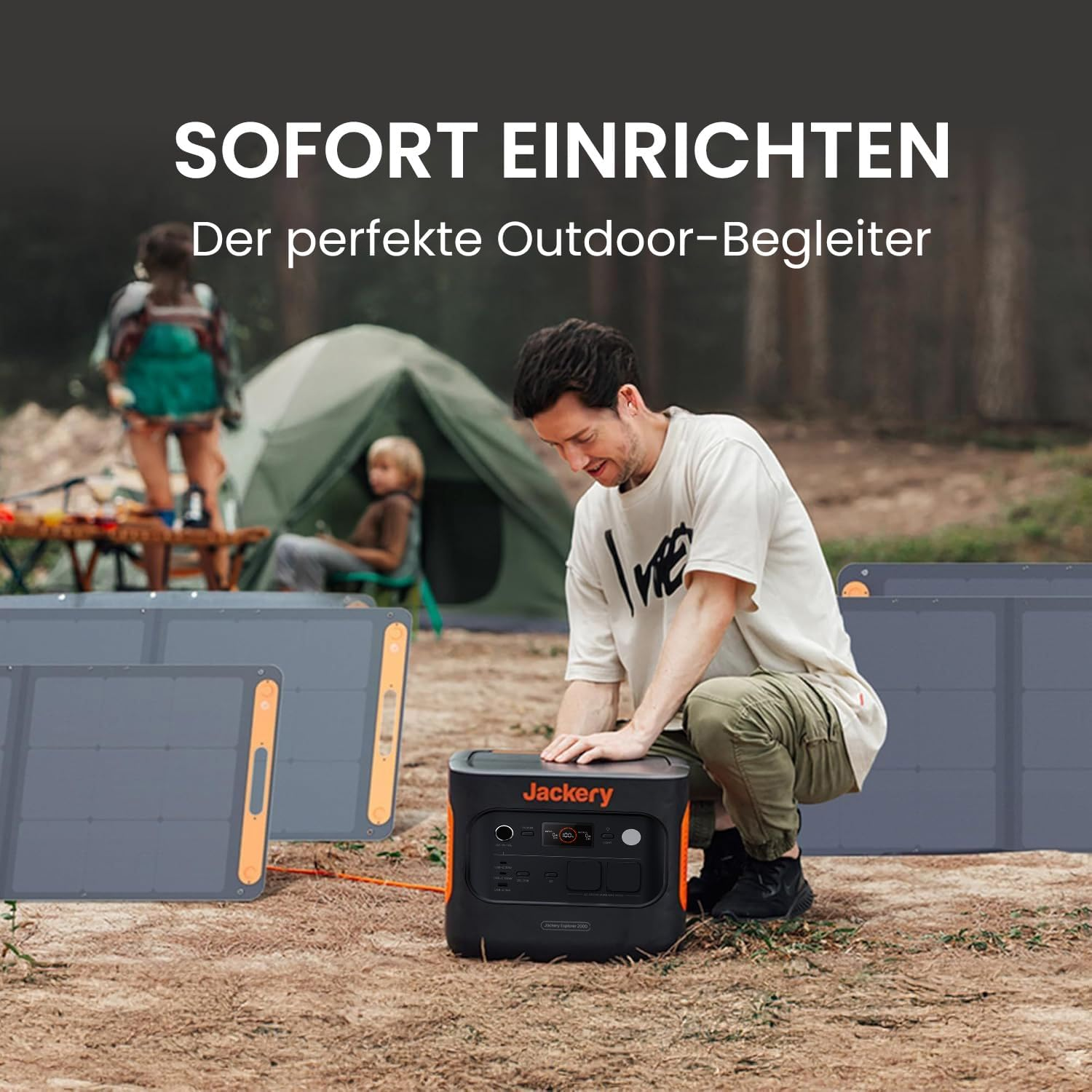 Mann richtet Jackery-Gerät im Freien ein, mit Solarpanelen und Zelt im Hintergrund. Familie beim Camping im Wald.