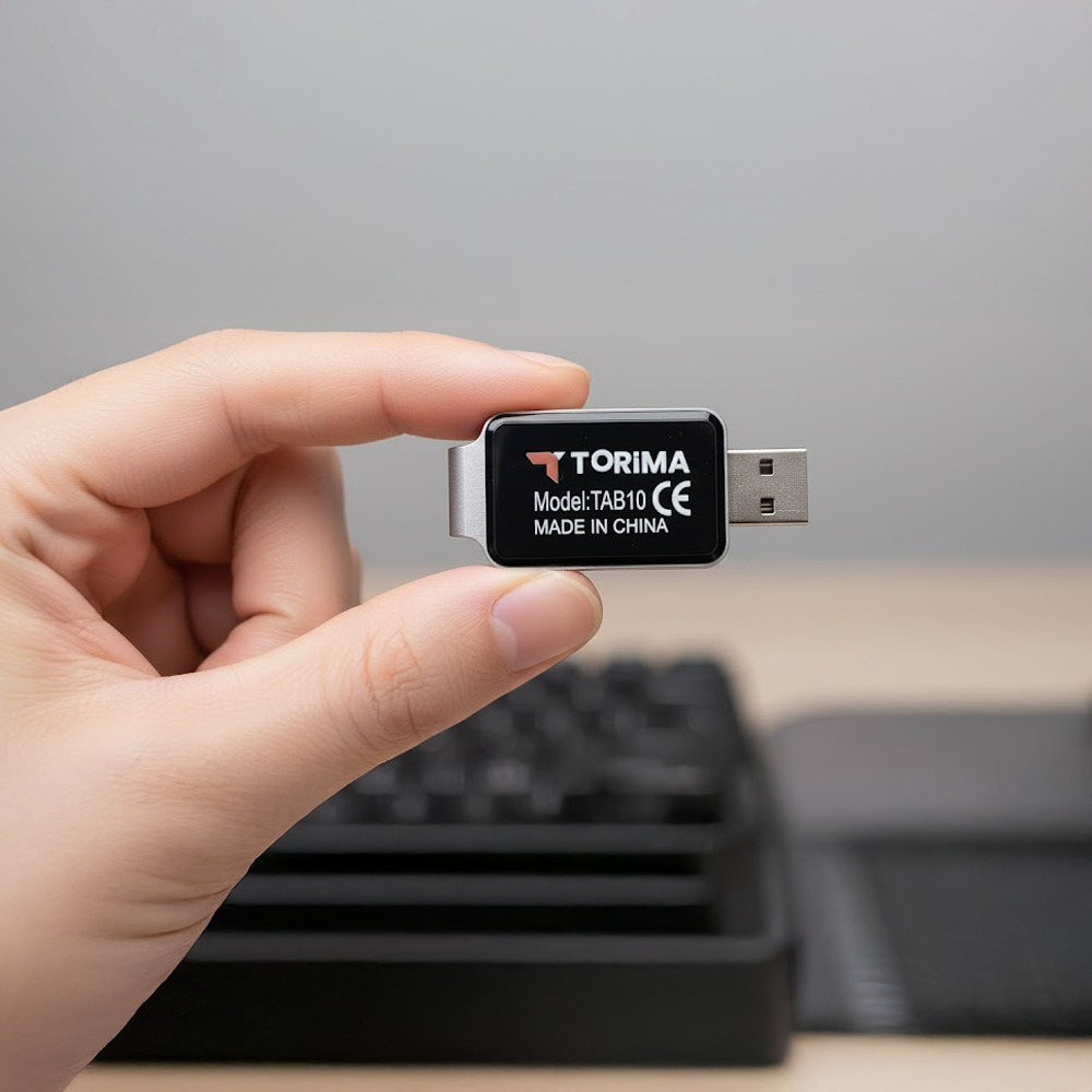 Bir el bir USB sürücüsü tutuyor. Sürücü, siyah, gümüş kenarlı ve Torima markalı. Arka planda bir klavye görülüyor.