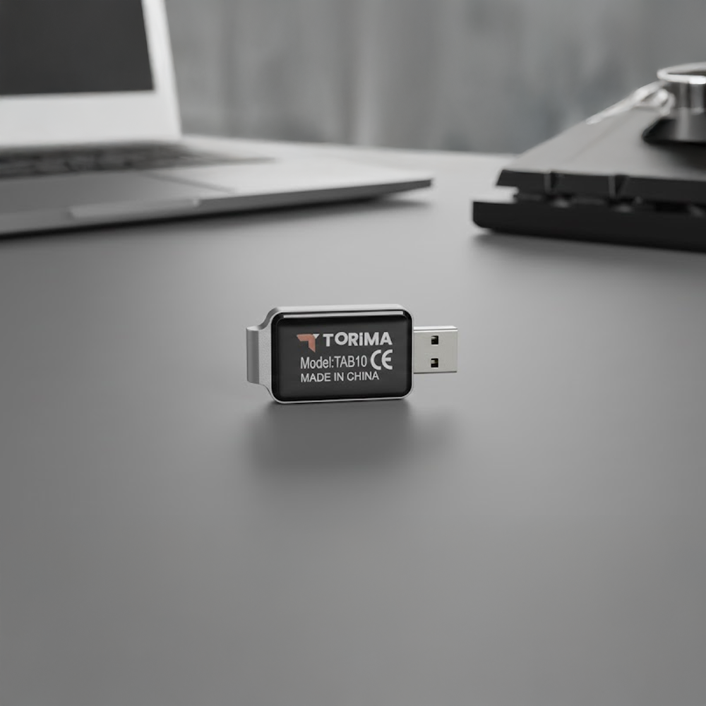 'TORIMA' etiketli bir USB sürücü gri bir yüzeyde duruyor. Bulanık arka planda bir dizüstü bilgisayar ve bir klavye var.
