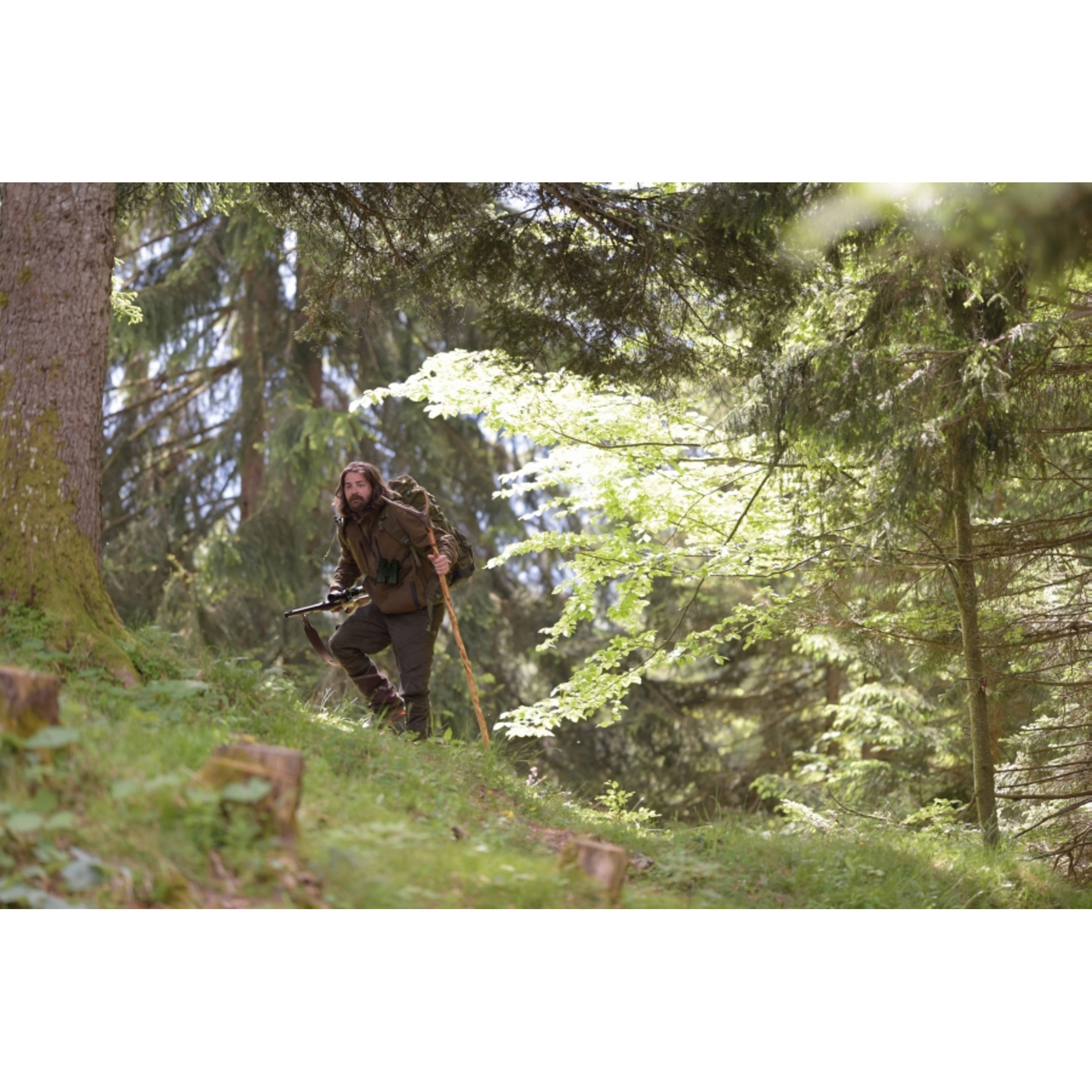 Un uomo cammina in una foresta, portando uno zaino, un fucile e un bastone. Gli alberi lo circondano su una collina verde.