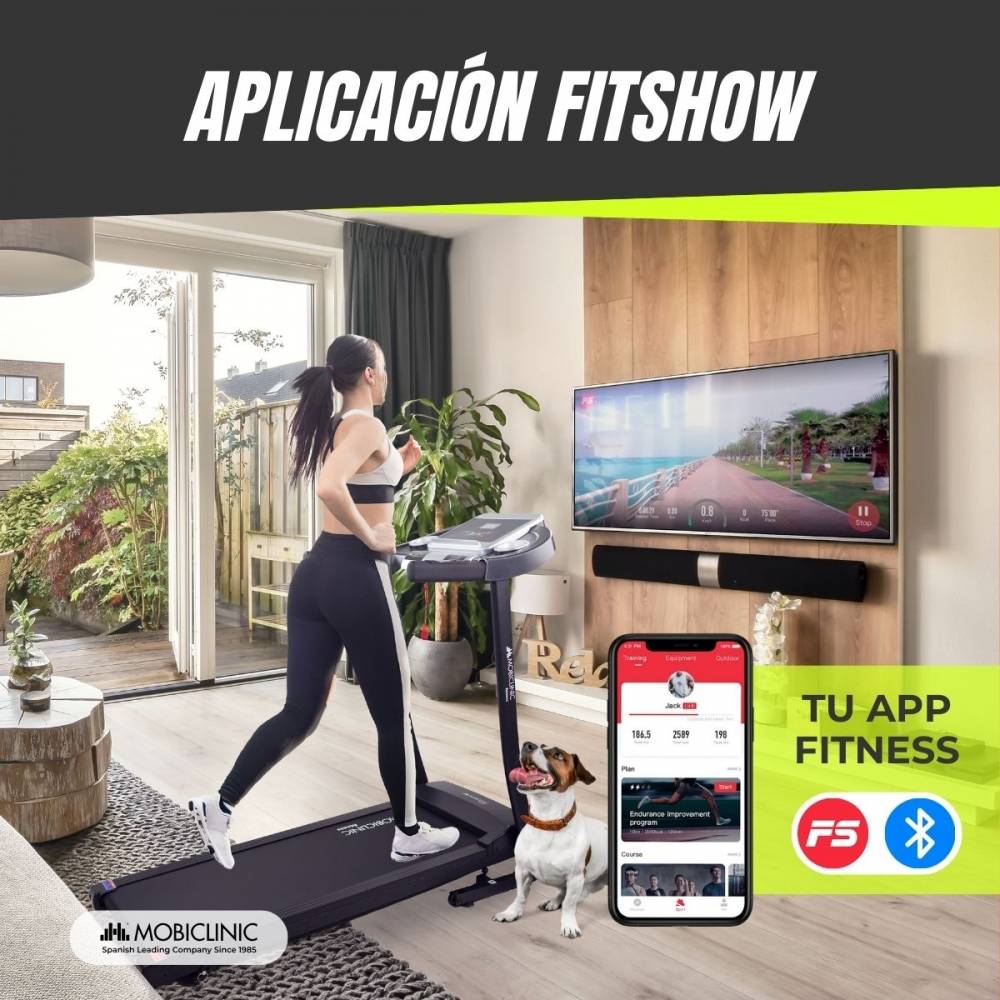Mujer corriendo en una cinta de correr en casa, pantalla de TV mostrando una pista de atletismo, perro sentado junto a la cinta, teléfono con una app de fitness.