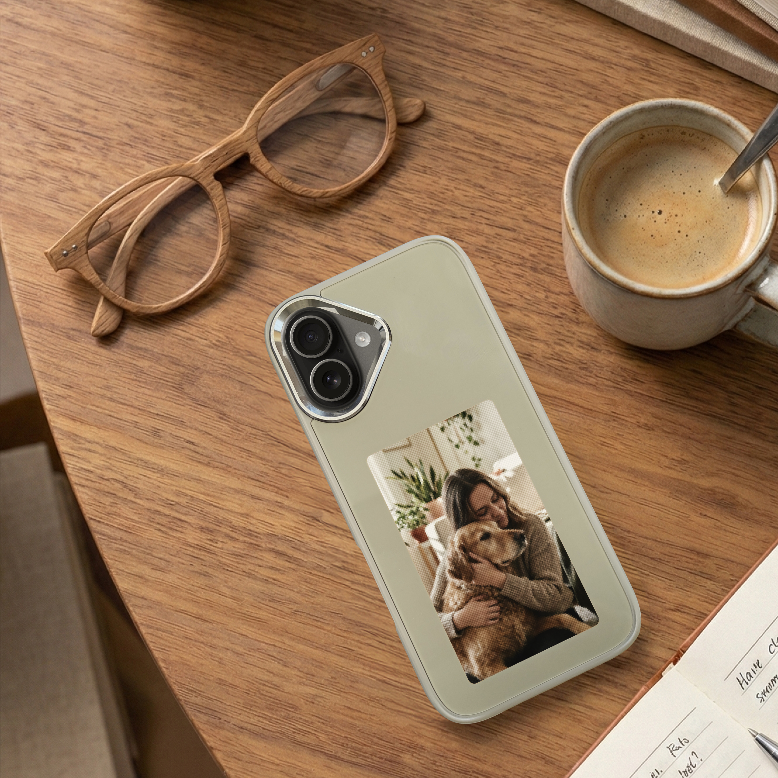 Houten tafel met telefoonhoesje met een vrouw die een hond omhelst, een bril, koffie en een notitieboekje.