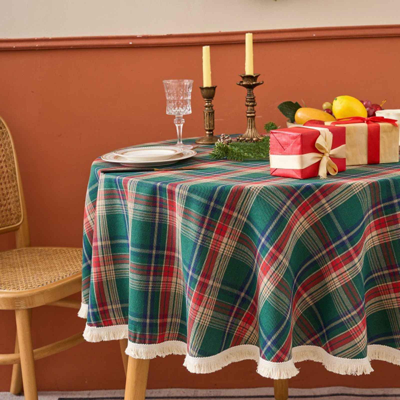 Ein runder Tisch mit einer grün-rot karierten Tischdecke ist für eine Mahlzeit gedeckt. Geschenke, Kerzen und Obst sind darauf.
