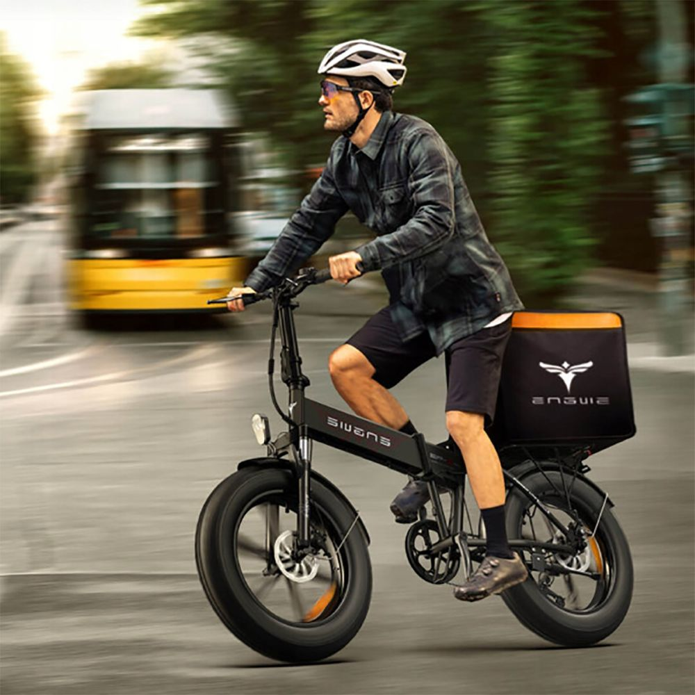 Un uomo guida una bicicletta elettrica nera con un baule. Indossa un casco e occhiali da sole. Un autobus giallo è sullo sfondo.