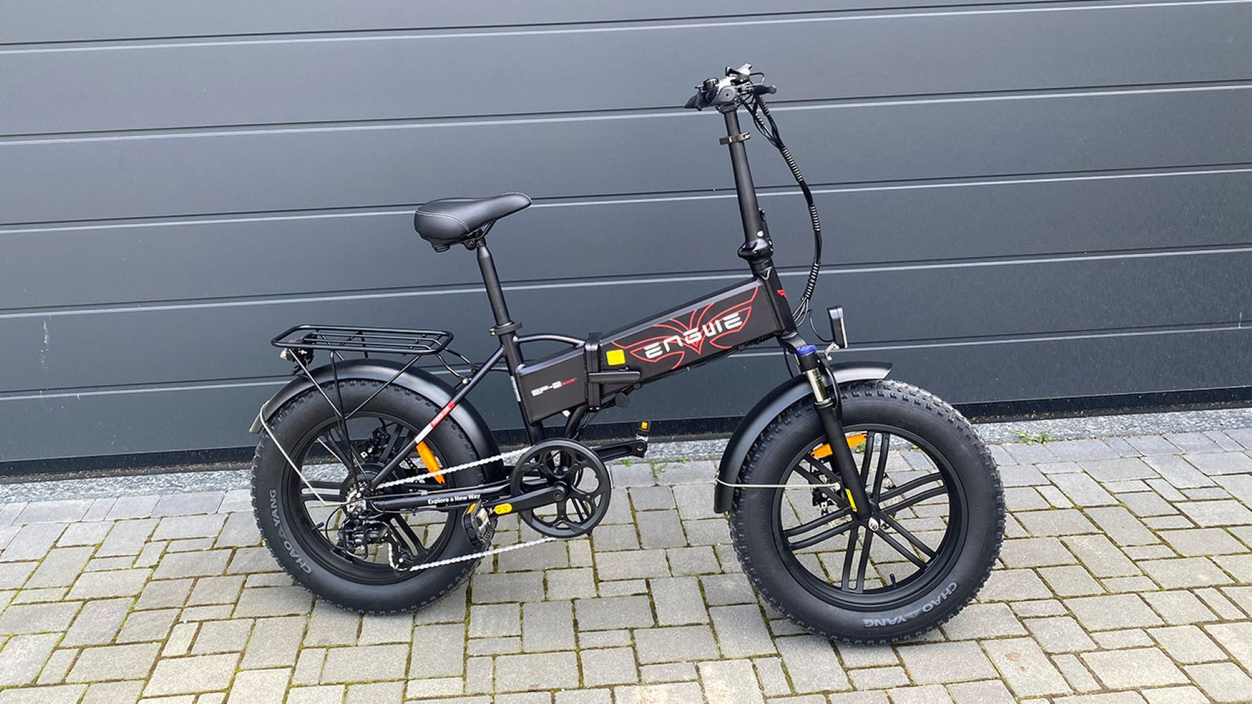 Bicicleta eléctrica negra estacionada sobre pavimento de ladrillo. La bici tiene llantas gordas, un portaequipajes y la marca 'ENGWE'.