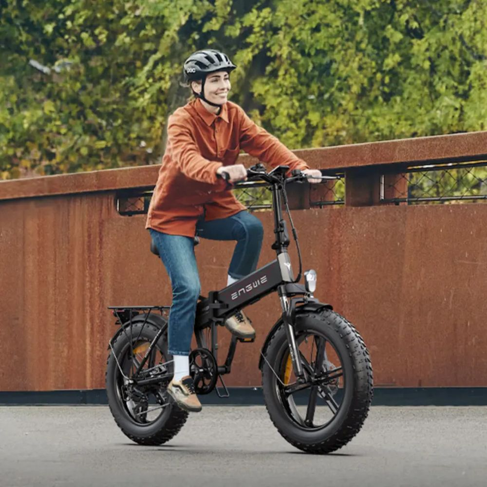 Una donna sorride mentre guida una e-bike ENGWE nera su una strada asfaltata. Indossa un casco, una camicia arancione e jeans blu.