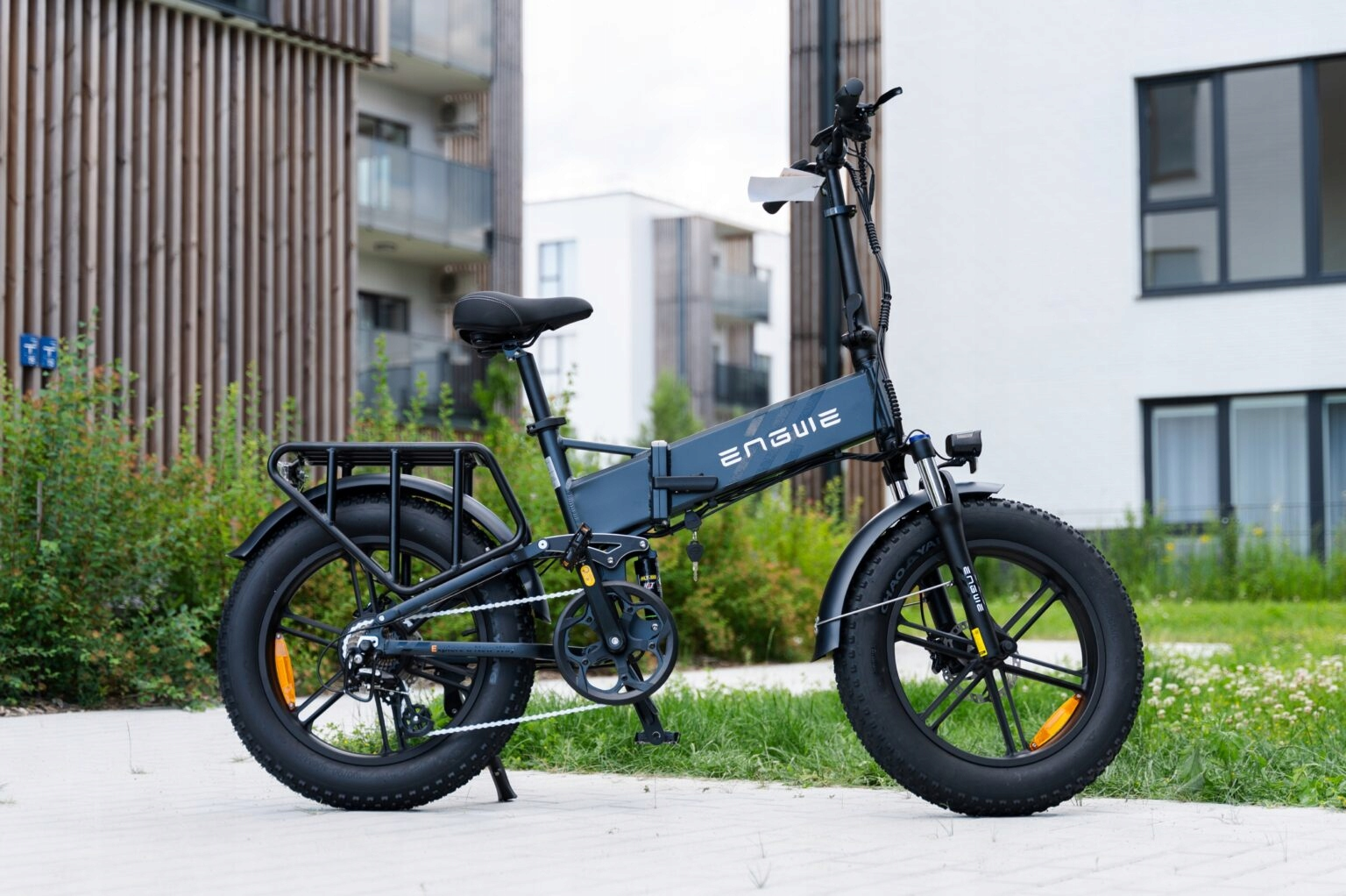 Una bicicleta eléctrica está estacionada al aire libre. Es negra y gris con un asiento negro. En el fondo hay edificios y vegetación.