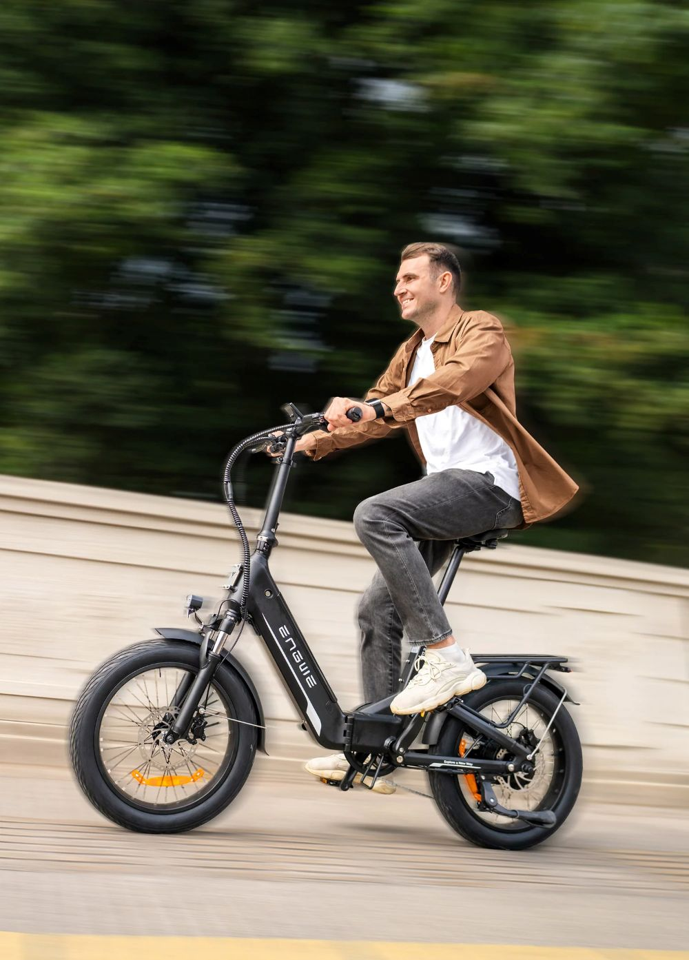 Un hombre monta una bicicleta eléctrica negra. Viste camisa marrón y pantalones grises. El fondo es verde y beige borroso.