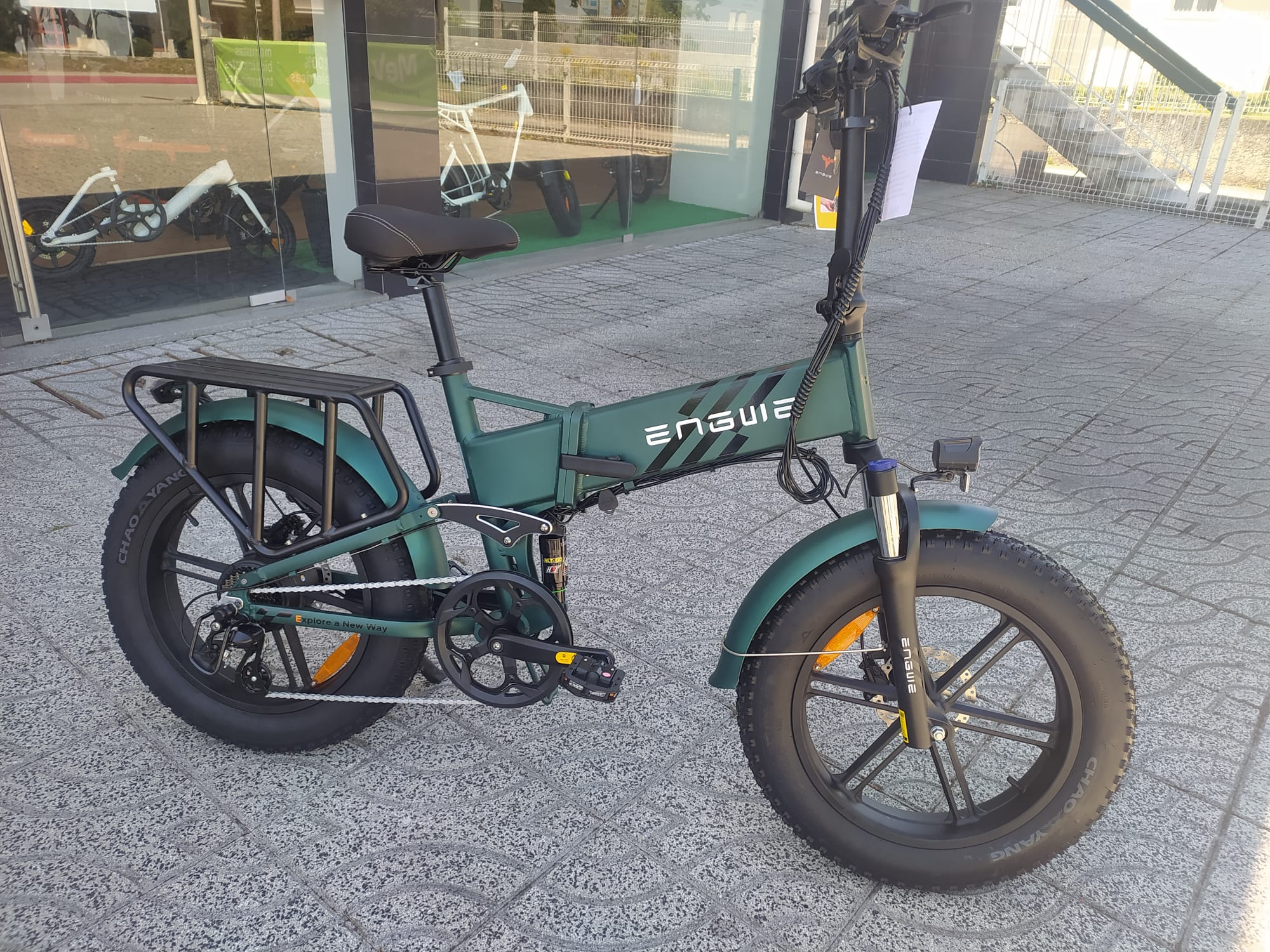Una e-bike ENGWE verde estacionada sobre una superficie embaldosada. Una tienda en el fondo.