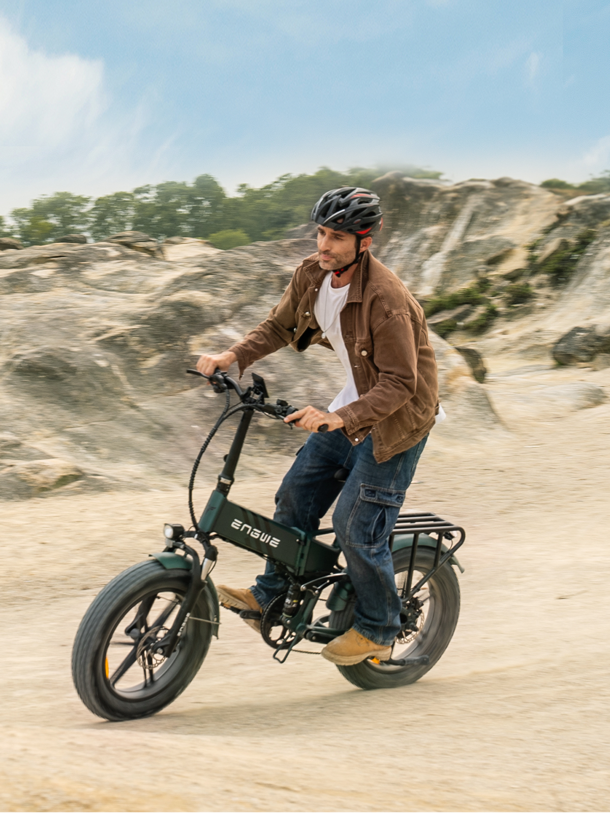 Ein Mann fährt mit einem E-Bike auf einem sandigen Weg. Er trägt eine braune Jacke, Jeans und einen Helm. Das Fahrrad ist dunkelgrün.