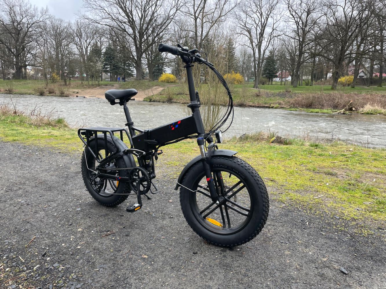 Bicicletta elettrica nera su un sentiero di ghiaia vicino a un fiume. Alberi sullo sfondo. La bici ha pneumatici grandi e un portapacchi.