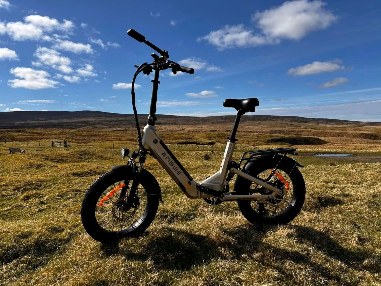 Una bici elettrica su una collina erbosa. Il cielo è blu con nuvole. La bici è bianca e nera.