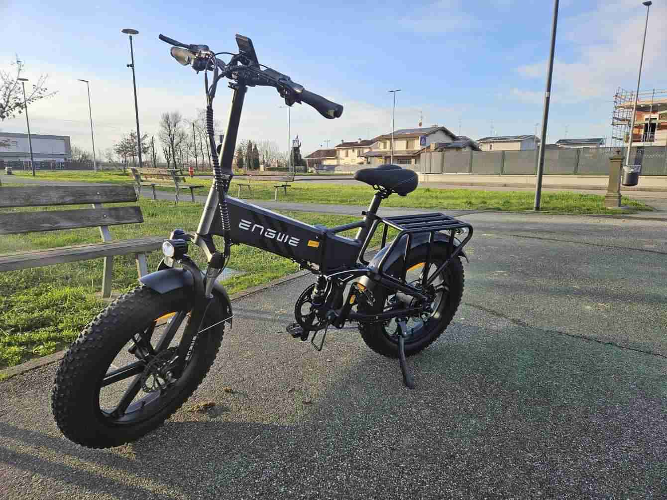 Una e-bike Engwe negra estacionada en un área pavimentada. La bicicleta tiene llantas anchas, portaequipajes y batería visible. Casas y cielo al fondo.
