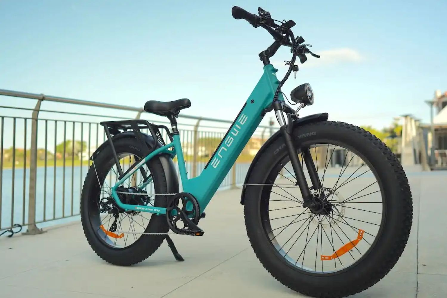 Una bicicleta eléctrica ENSURE turquesa sobre un camino de hormigón, cerca del agua y una barandilla metálica. Cielo despejado al fondo.