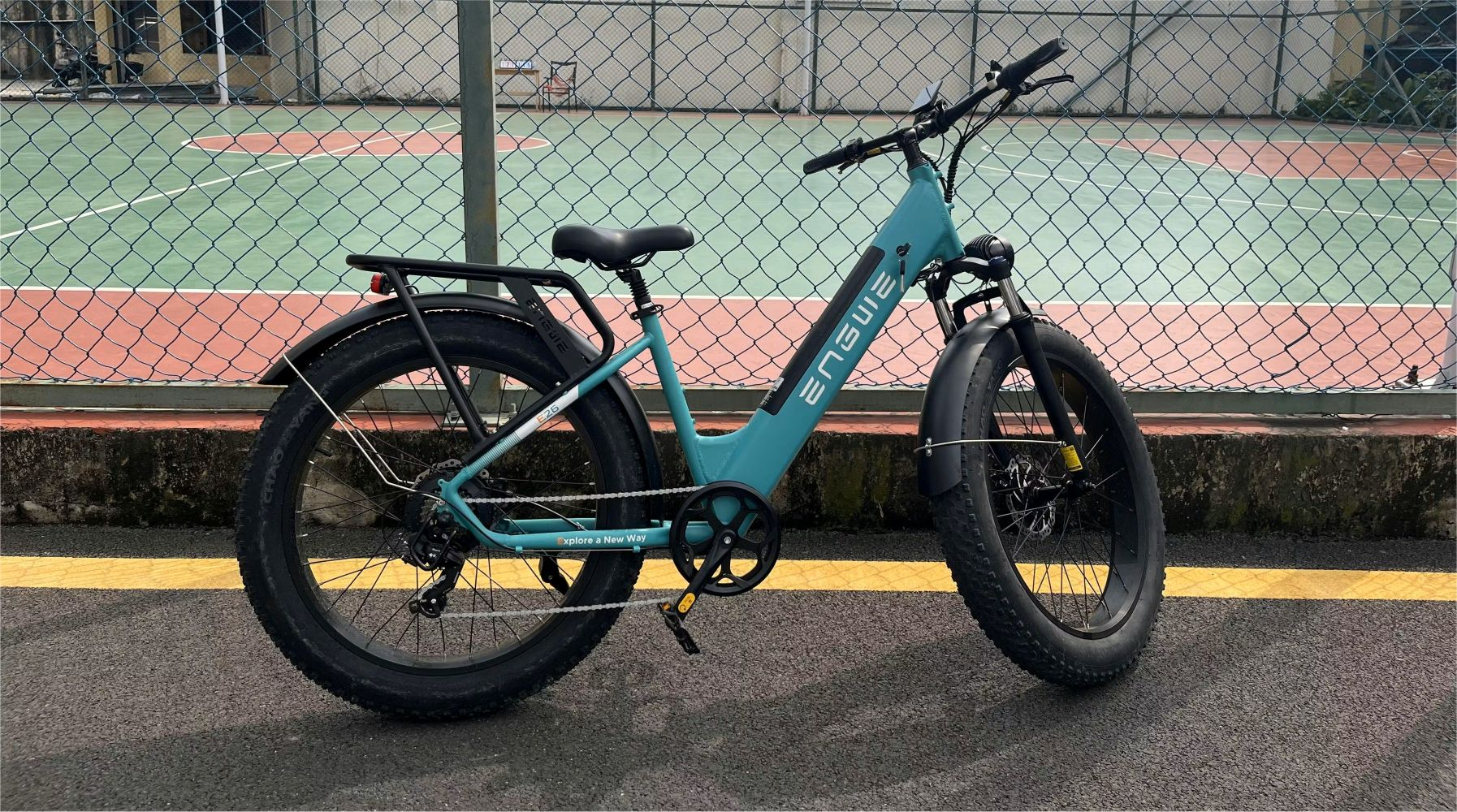 Una bicicleta eléctrica turquesa estacionada en asfalto. Una cancha de tenis está al fondo.