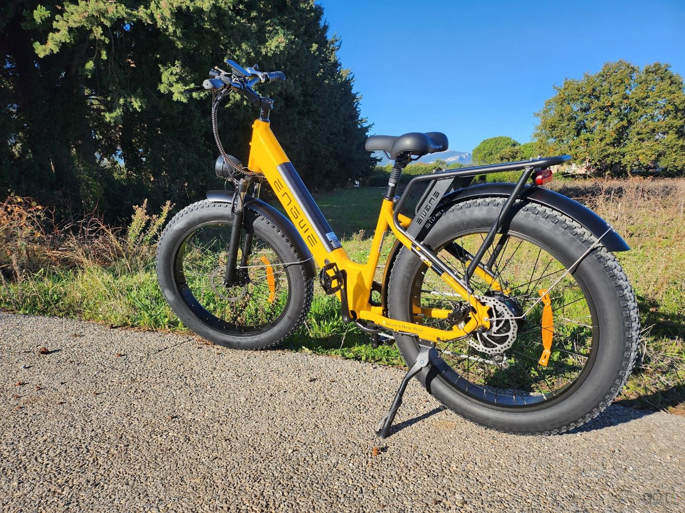 Una bicicleta eléctrica Engwe amarilla en un camino. Llantas, asiento y portaequipajes negros. Árboles y cielo azul al fondo.