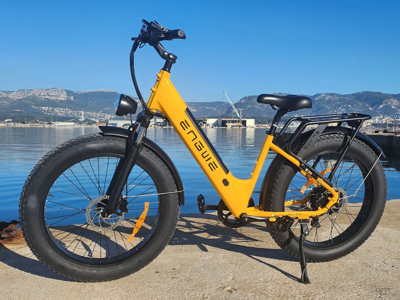 Bicicleta eléctrica Engwe amarilla estacionada en concreto, con neumáticos negros, contra un mar azul, montañas y cielo.