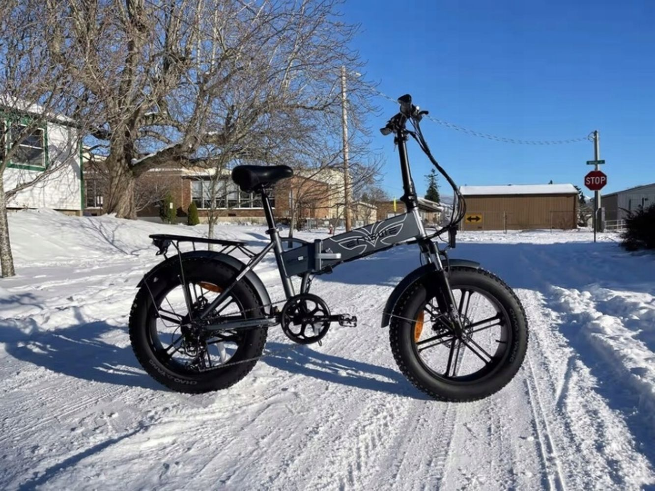 Bicicletta elettrica su strada innevata. Edifici e alberi sullo sfondo, cielo blu.