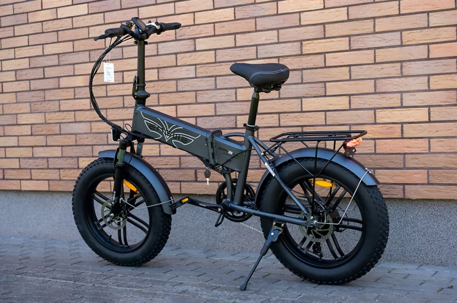 Una bicicletta elettrica nera è parcheggiata di fronte a un muro di mattoni. Ha pneumatici grandi e un portapacchi. La bici è su un pavimento di mattoni.