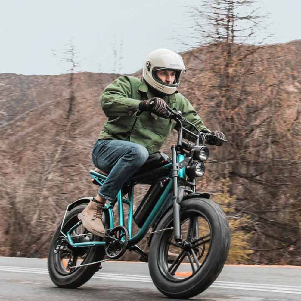 Un hombre monta una bicicleta eléctrica en una carretera. Lleva casco, chaqueta verde y vaqueros. La bicicleta es turquesa y negra.
