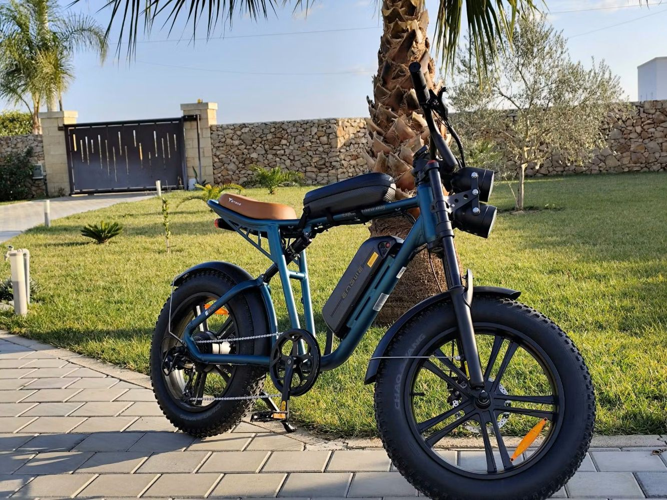 Una bicicleta eléctrica está estacionada en un camino de ladrillos, con césped y un muro de piedra al fondo.