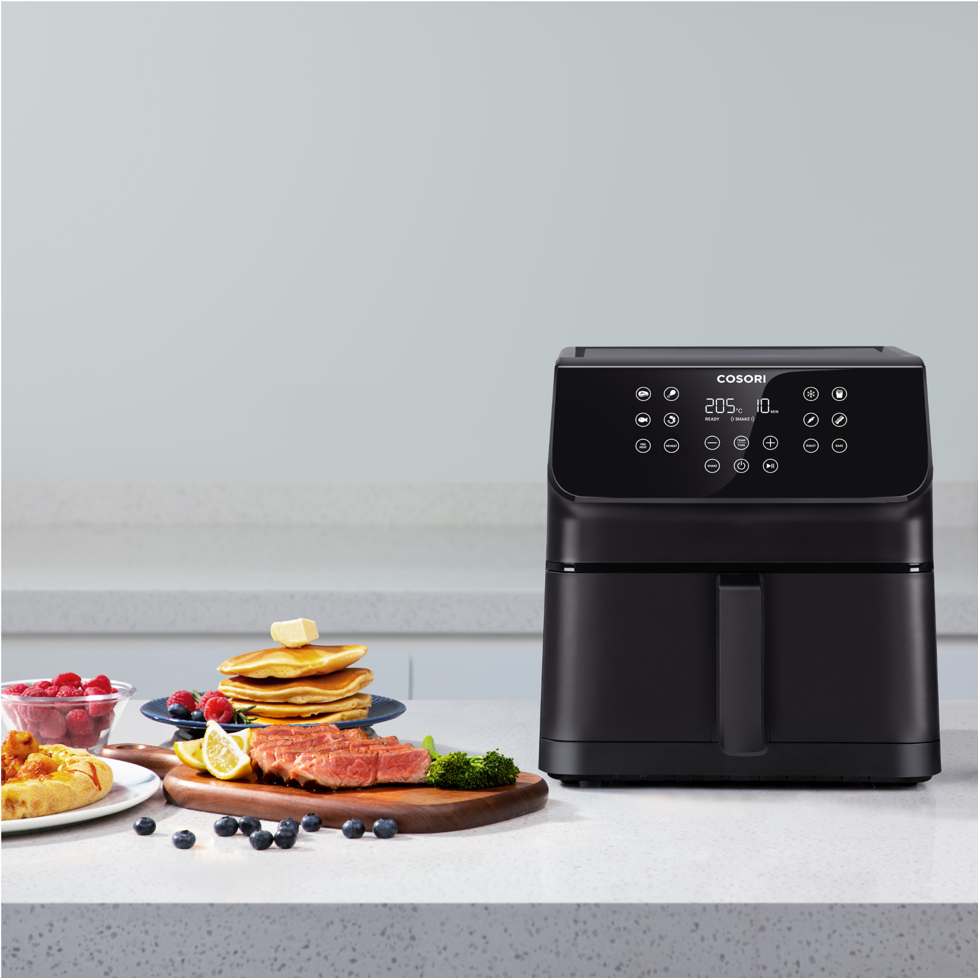 Een zwarte Cosori airfryer staat naast een ontbijt met pannenkoeken, biefstuk en bessen op een wit aanrecht.