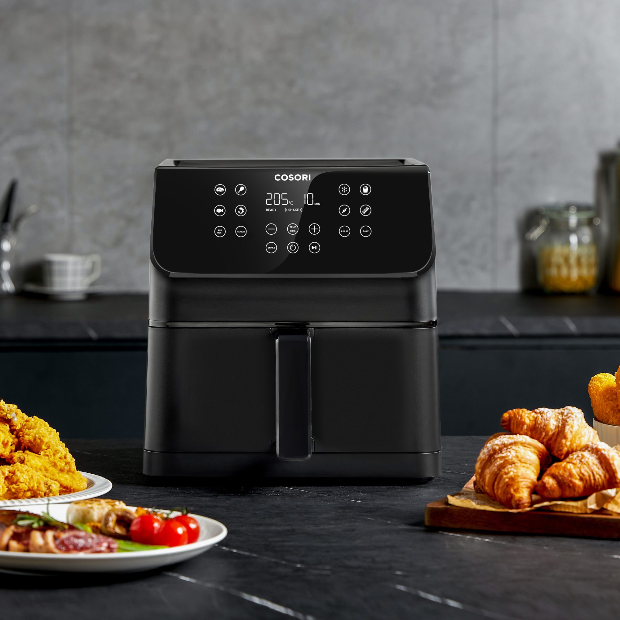 Een zwarte airfryer met eten. Croissants, gefrituurde kip en tomaten op tafel. Het display toont 205 graden.