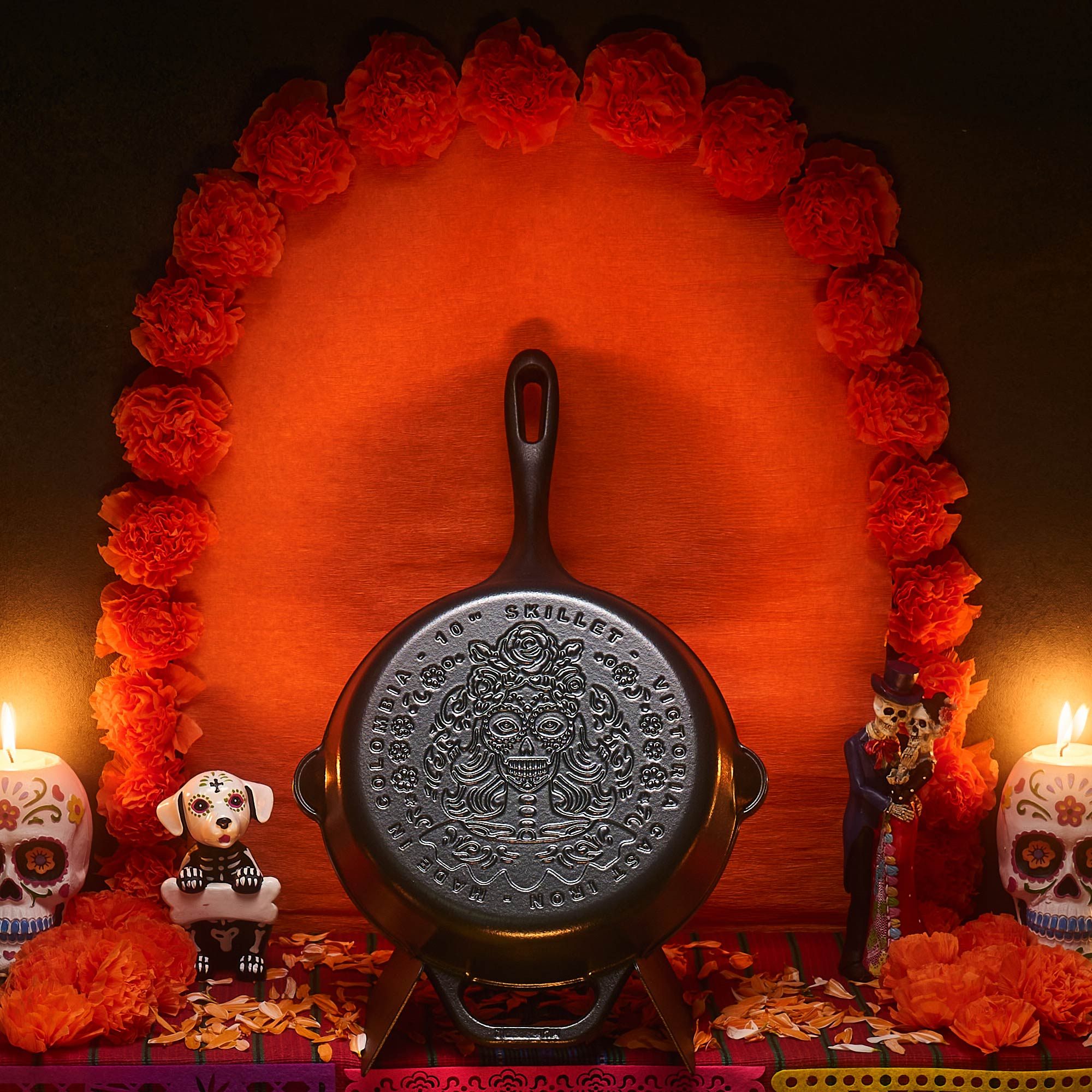 Eine Dia de los Muertos-Anzeige mit einer Bratpfanne, orangefarbenen Blumen, Kerzen und Skelettfiguren auf einem rot-orangen Hintergrund.