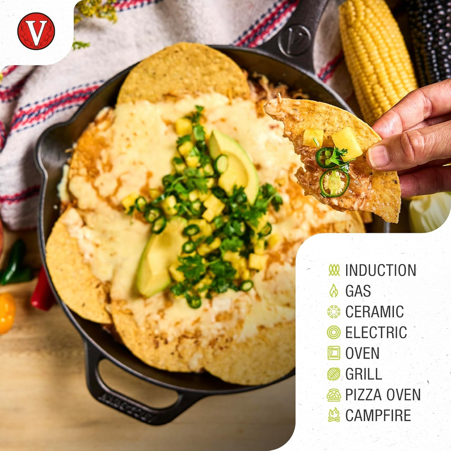 Eine gusseiserne Pfanne mit Nachos, Käse, Avocado und Belägen. Eine Hand hält einen Chip mit Belägen. Maiskolben im Hintergrund.
