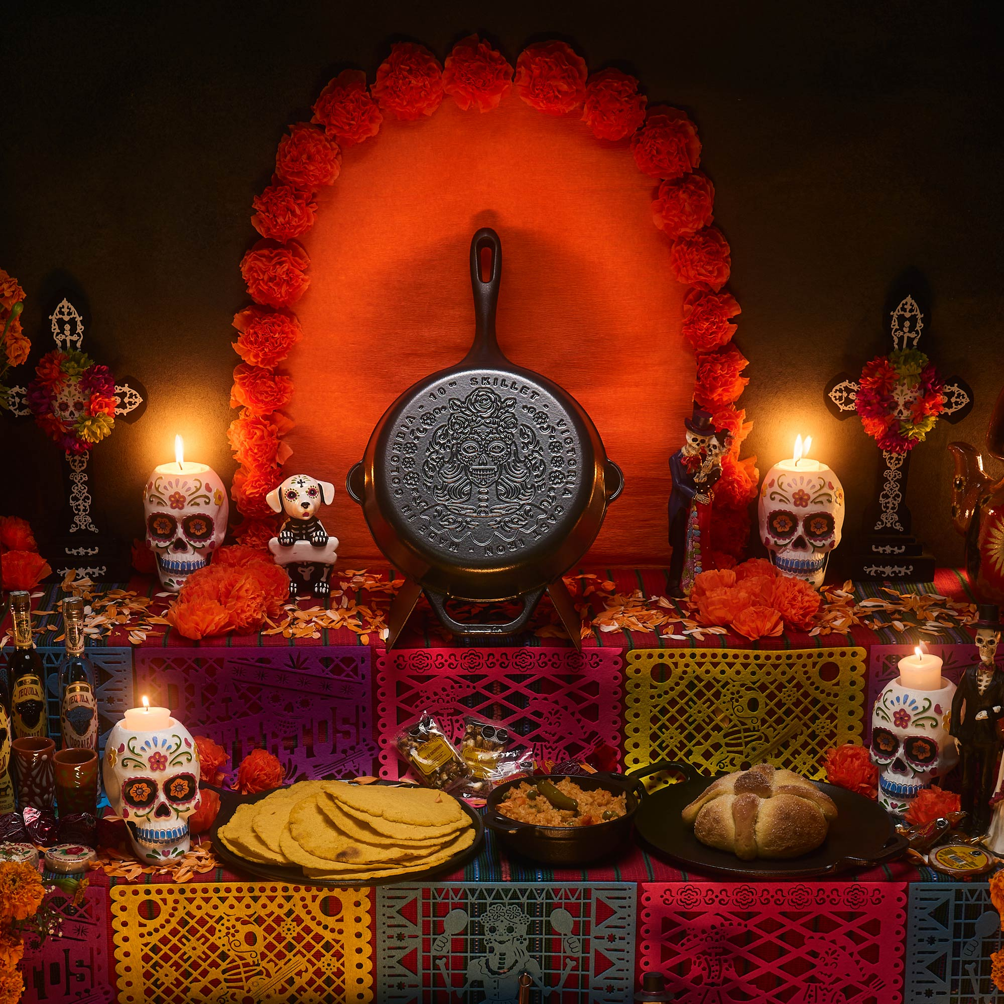 Dia de los Muertos-Altar mit Pfanne, Kerzen, Totenköpfen, Essen und Dekorationen auf bunt gemusterter Tischdecke.