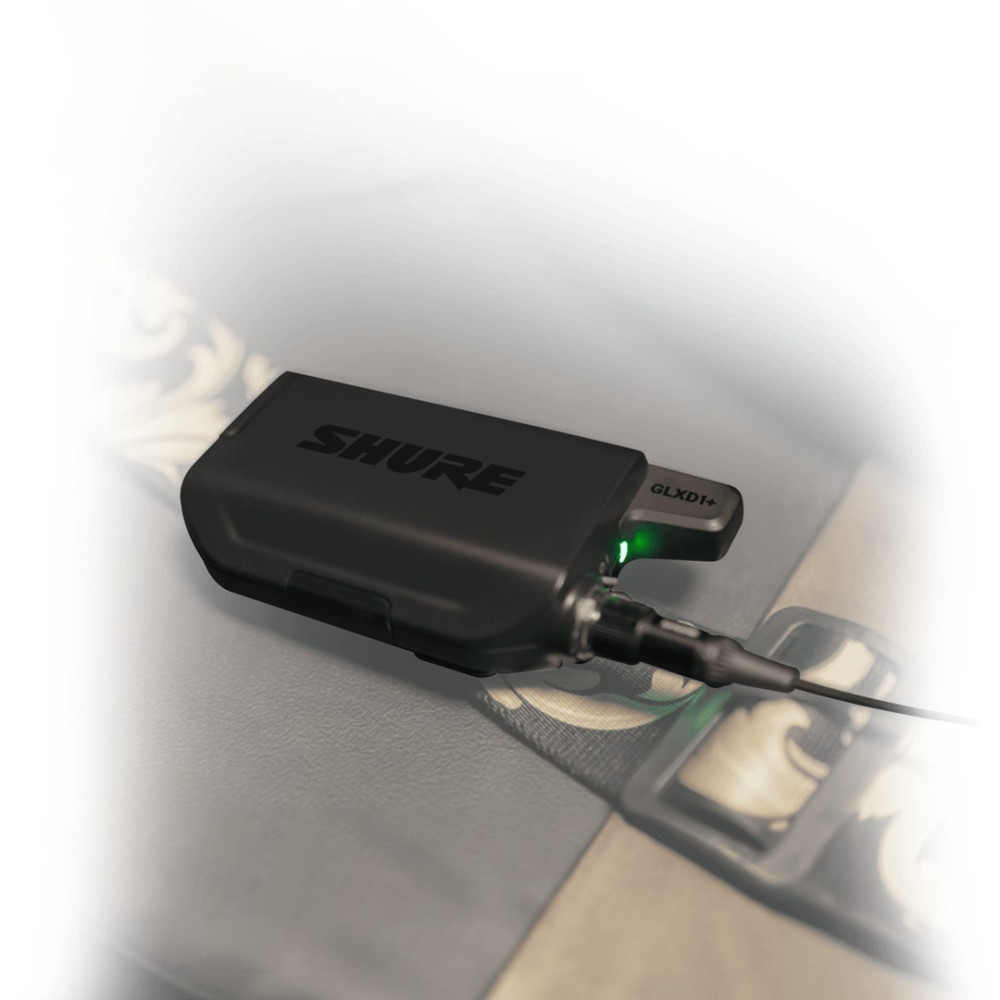 Czarny nadajnik bezprzewodowy Shure GLXD1+ leży na powierzchni. Świeci się zielone światło. Kable wychodzą z urządzenia.