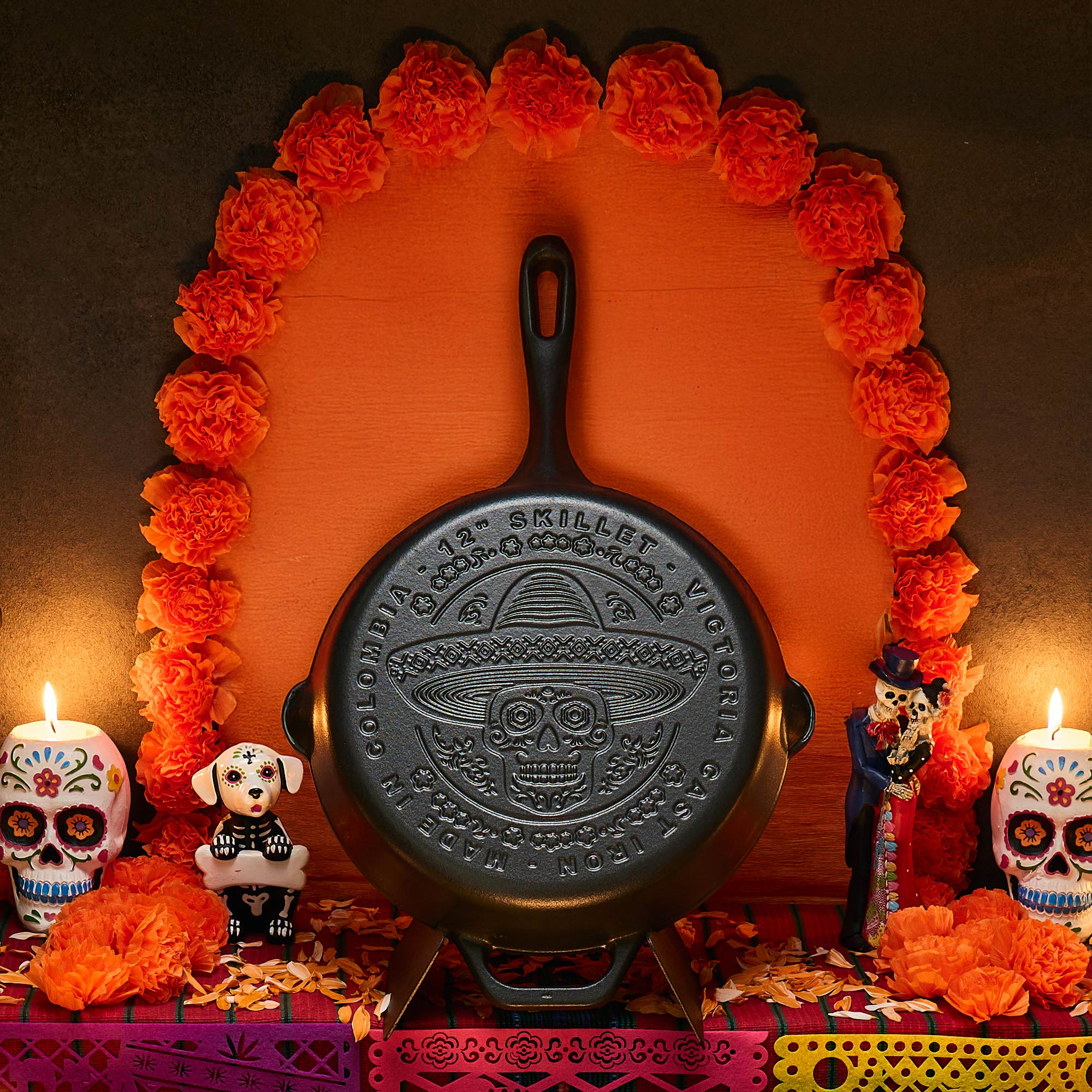 Eine gusseiserne Bratpfanne mit Totenkopfmotiv steht im Mittelpunkt, umgeben von orangefarbenen Blumen, Kerzen und Day-of-the-Dead-Figuren.