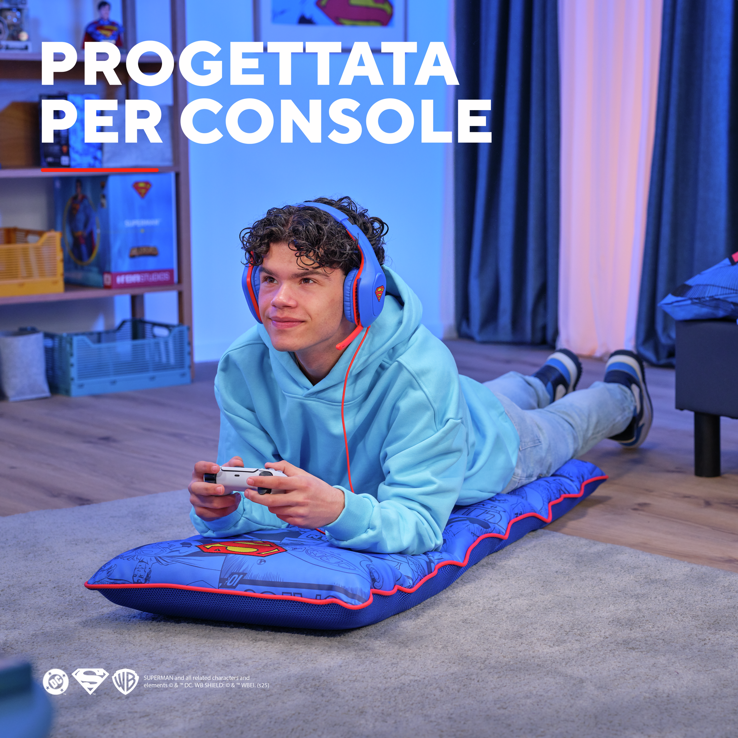 Un giovane è sdraiato su un tappeto di Superman, indossa le cuffie e tiene in mano un controller. Lo sfondo è blu.