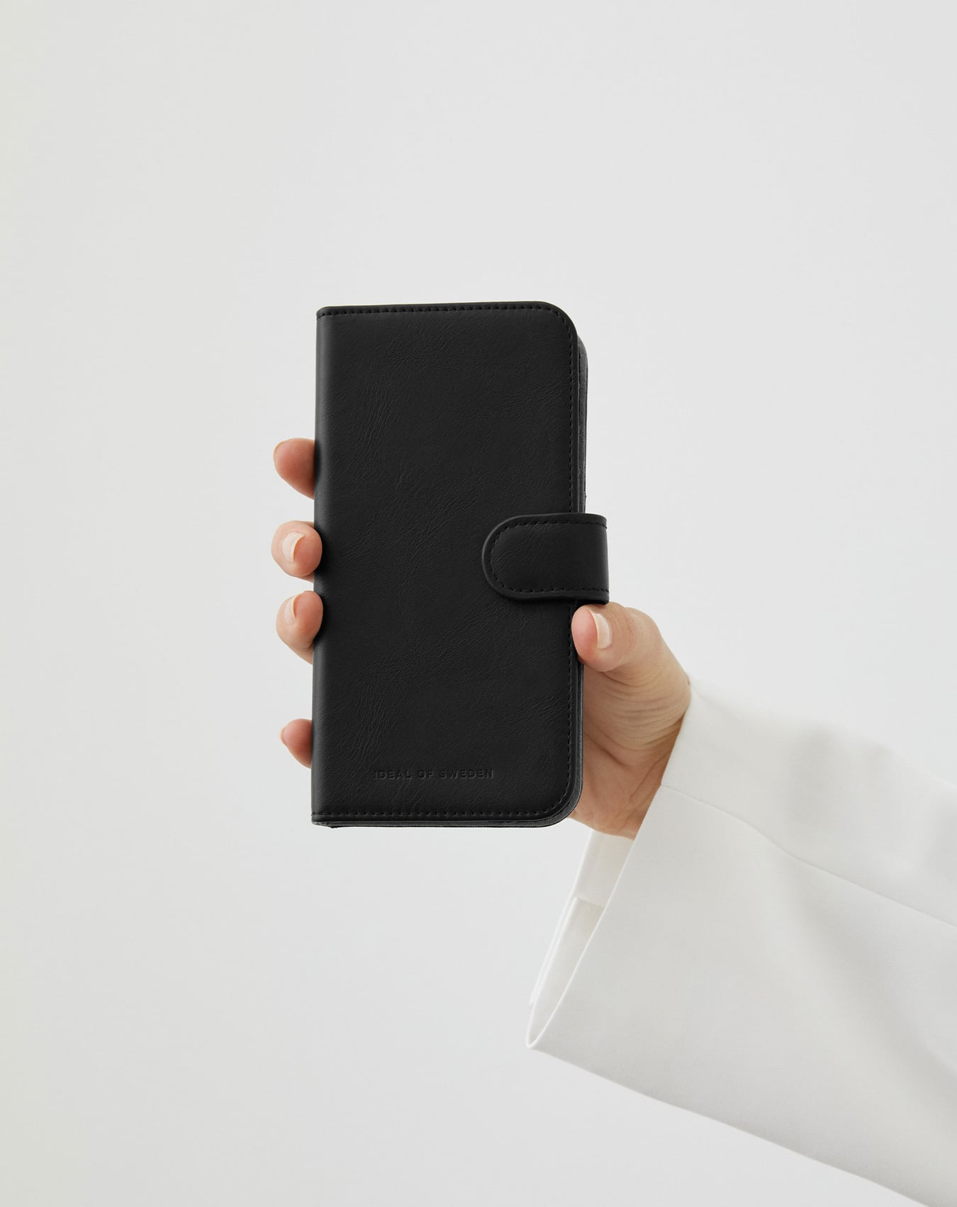 Eine Hand hält eine schwarze Handyhülle mit Magnetverschluss. Auf der Hülle steht 'IDEAL OF SWEDEN'. Der Hintergrund ist weiß.