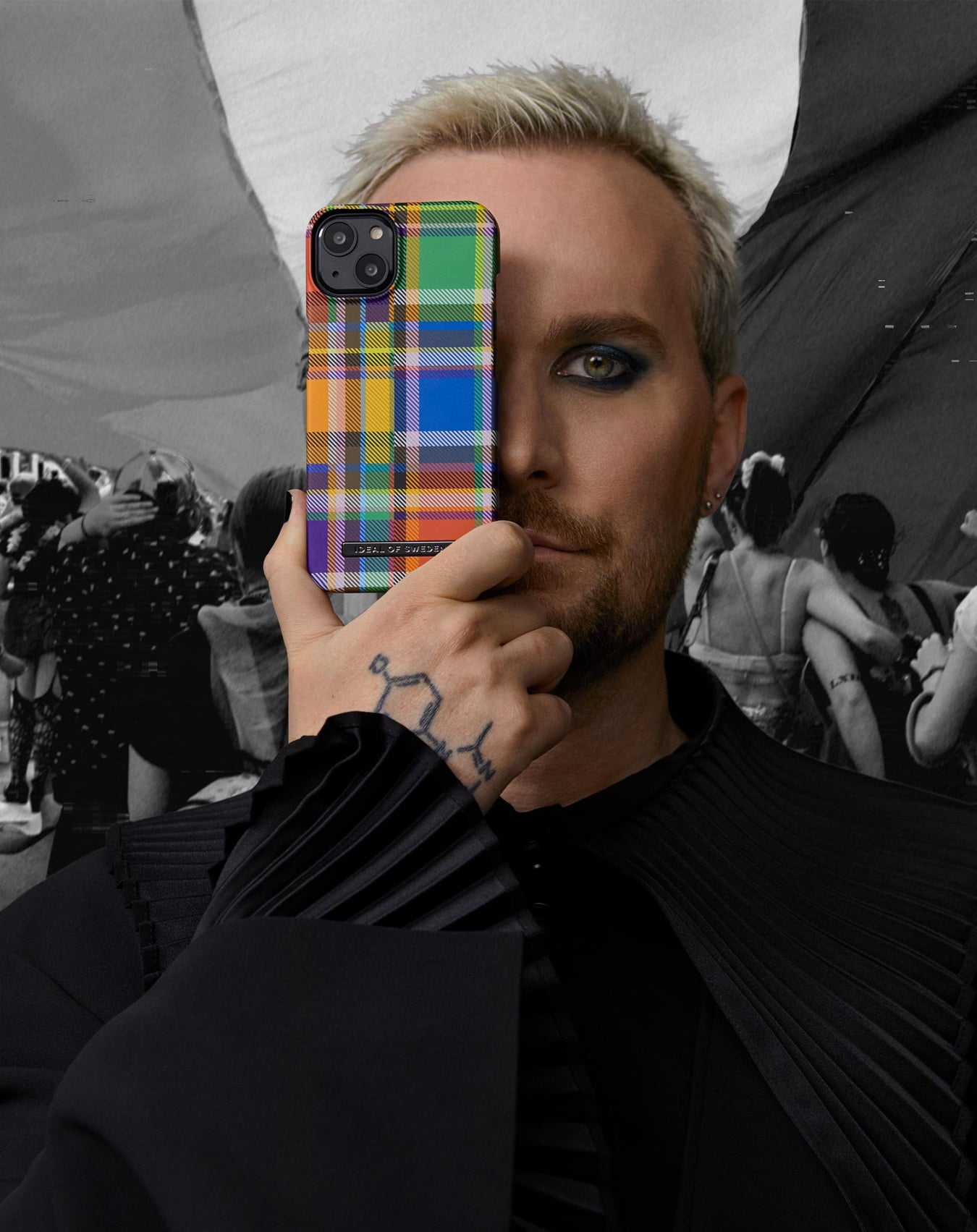 Ein Mann hält eine bunte karierte Handyhülle vor sein Gesicht. Im Hintergrund sind Menschen. Er hat Tätowierungen und dunkles Augen-Make-up.