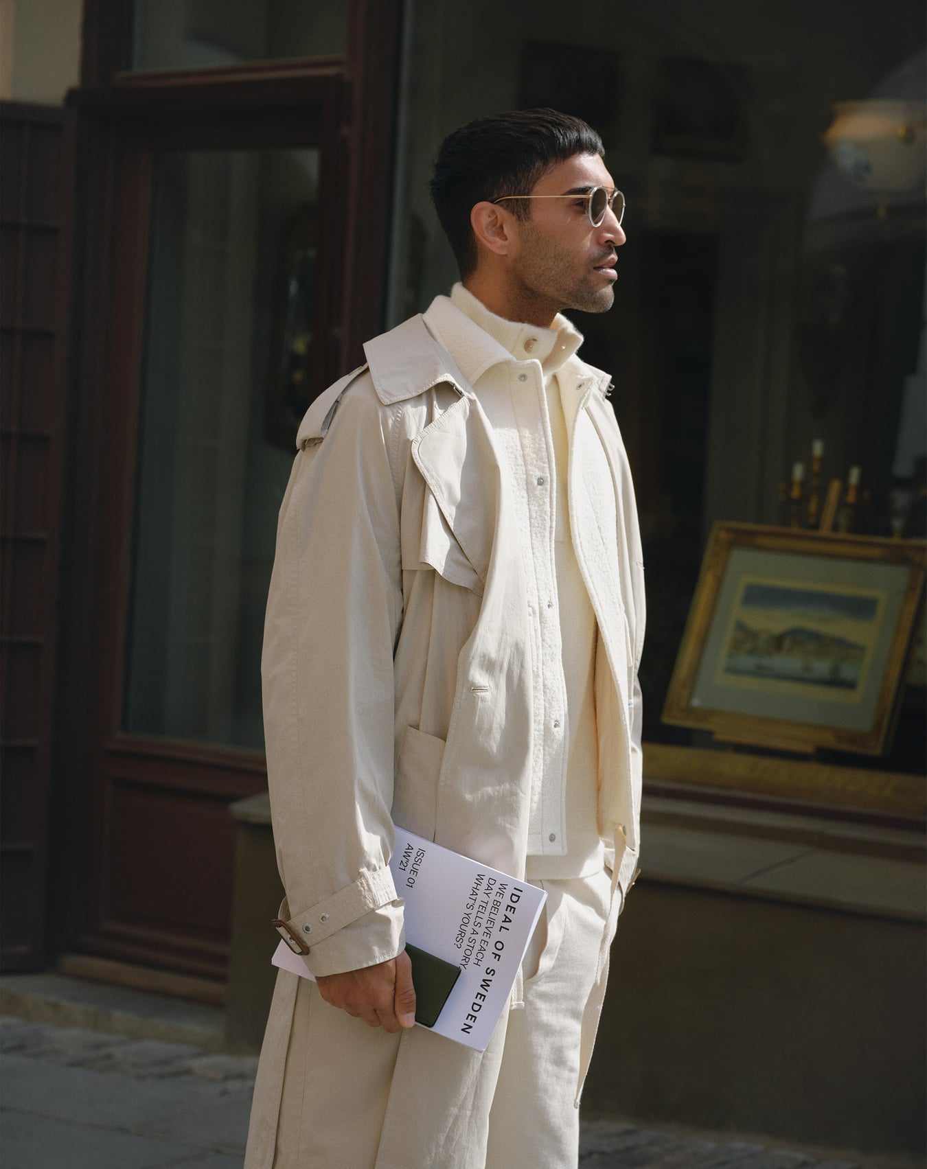 Ein Mann in cremefarbener Kleidung, Trenchcoat und Sonnenbrille steht draußen und hält ein Magazin mit dem Titel 'Ideal of Sweden'.