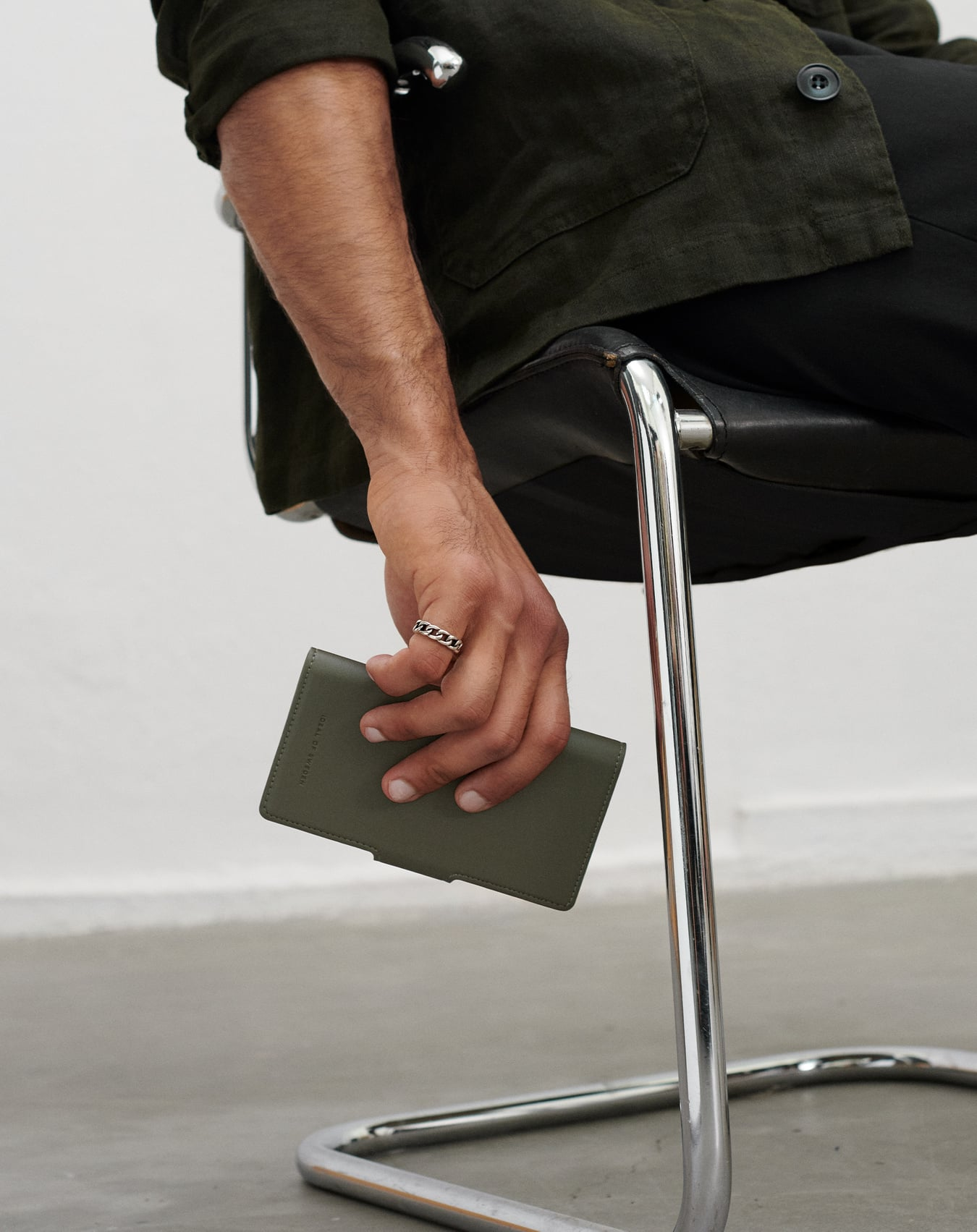 Ein Mann sitzt und hält eine grüne Brieftasche. Er trägt einen Ring. Er trägt eine dunkelgrüne Jacke und schwarze Hose.
