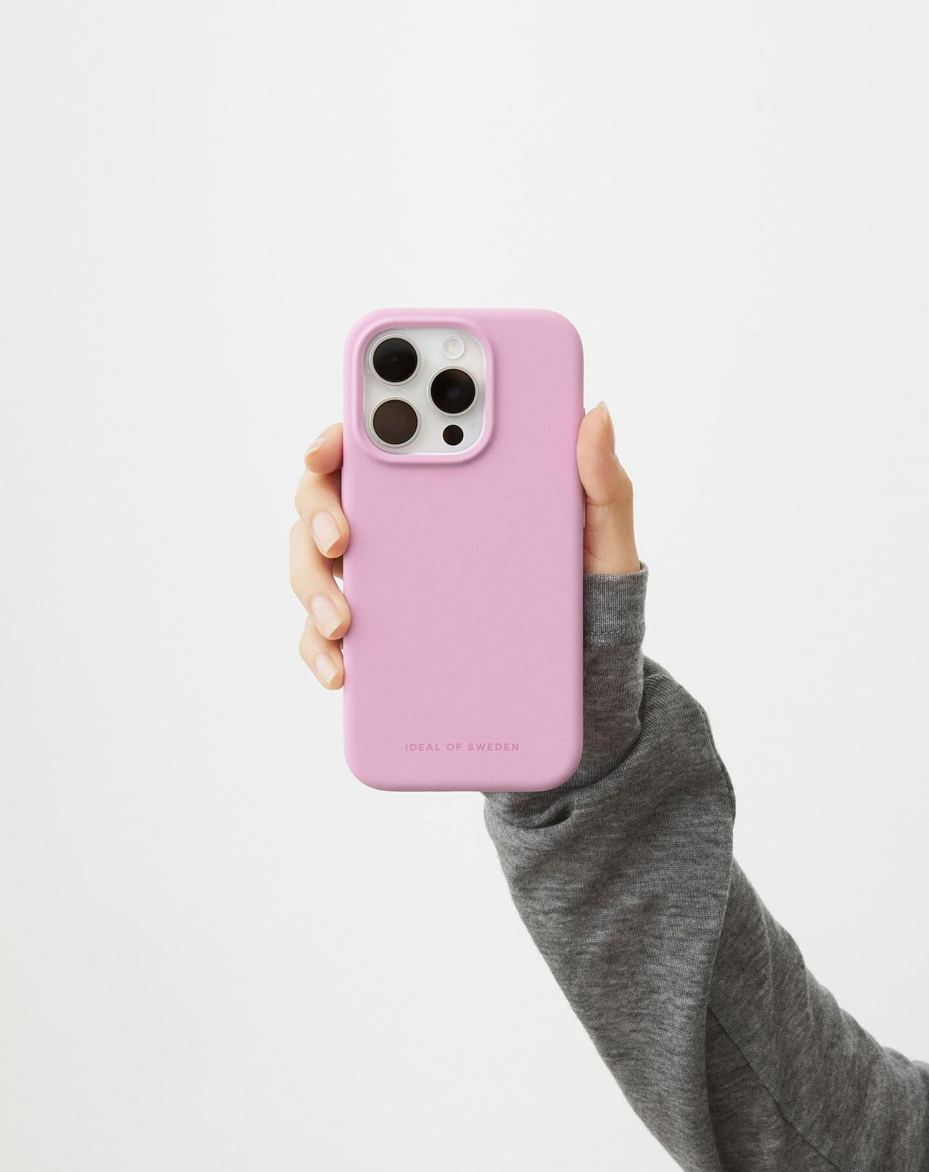 Eine Hand hält eine rosa Handyhülle mit Ideal of Sweden-Marke. Ein grauer Ärmel ist vor weißem Hintergrund sichtbar.