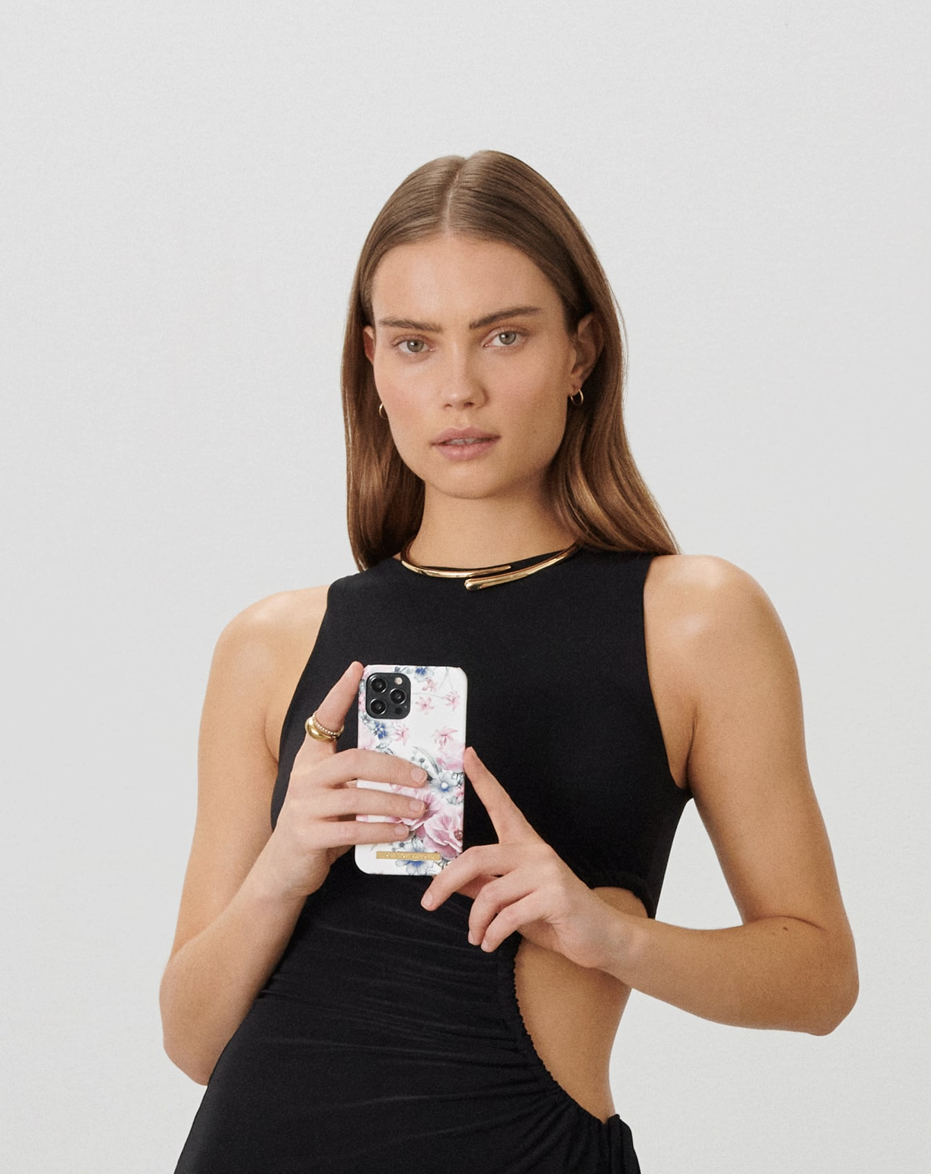 Frau in schwarzem Kleid hält ein Blumenhandy. Sie trägt Goldschmuck und blickt in die Kamera. Weißer Hintergrund.