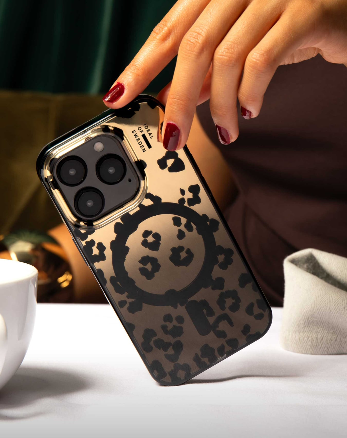 Eine Hand hält ein Telefon mit Leopardmuster-Hülle. Roter Nagellack. Weißer Tisch, weiße Tasse und Leinen-Serviette im Hintergrund.