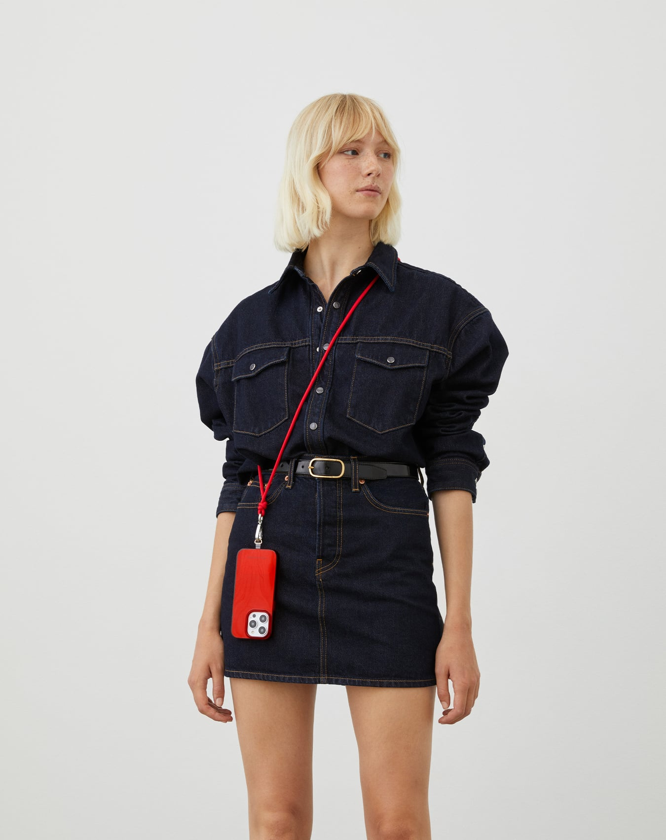 Eine Frau in Jeanshemd und -rock, die eine rote Handyhülle mit rotem Riemen hält. Sie trägt einen schwarzen Gürtel und hat blonde Haare.