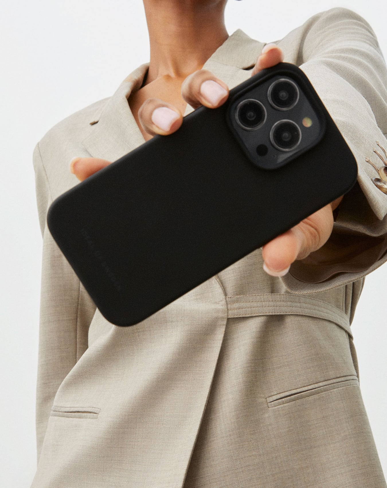 Eine Person hält ein schwarzes Telefon in einer Hülle und präsentiert die Kamera. Sie trägt einen beigen Blazer.
