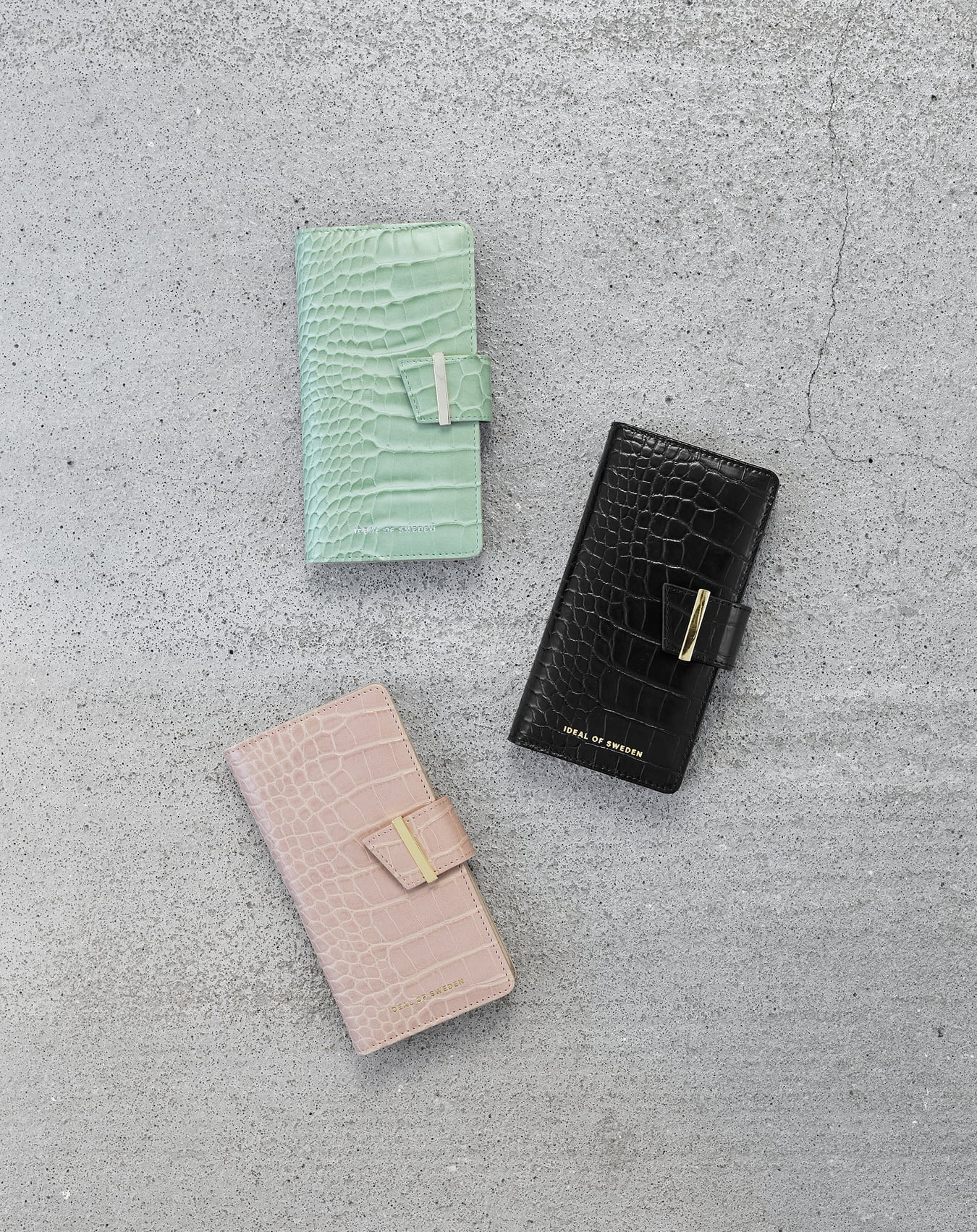 Drei Handyhüllen mit Krokodiltextur: Mint, Schwarz und Pink. Sie liegen auf einer grauen Betonoberfläche.