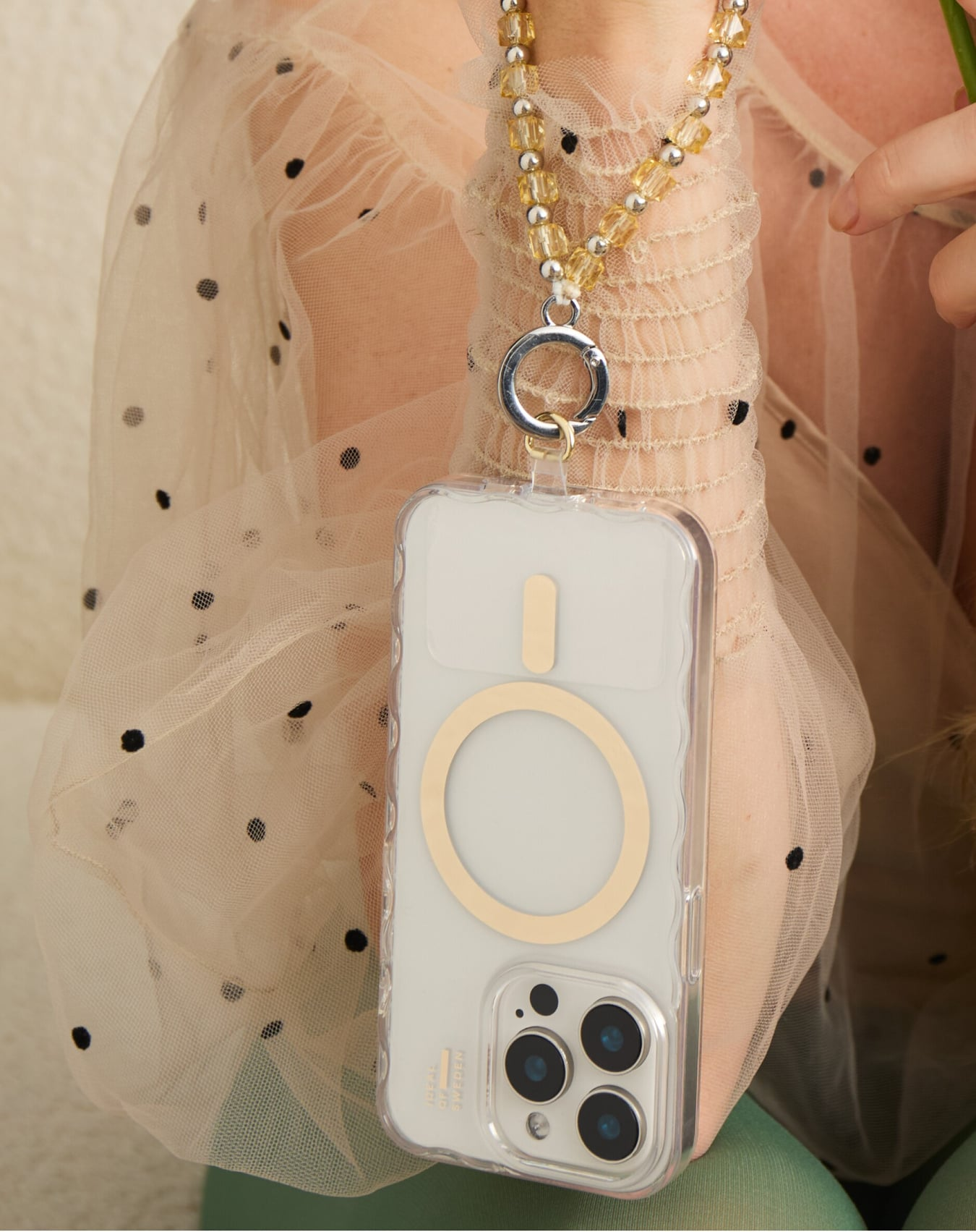 Eine Frau hält ein Telefon in einer transparenten Hülle, die an einer Perlenkette befestigt ist. Sie trägt ein durchsichtiges Oberteil mit Punkten. Der Hintergrund ist weich.