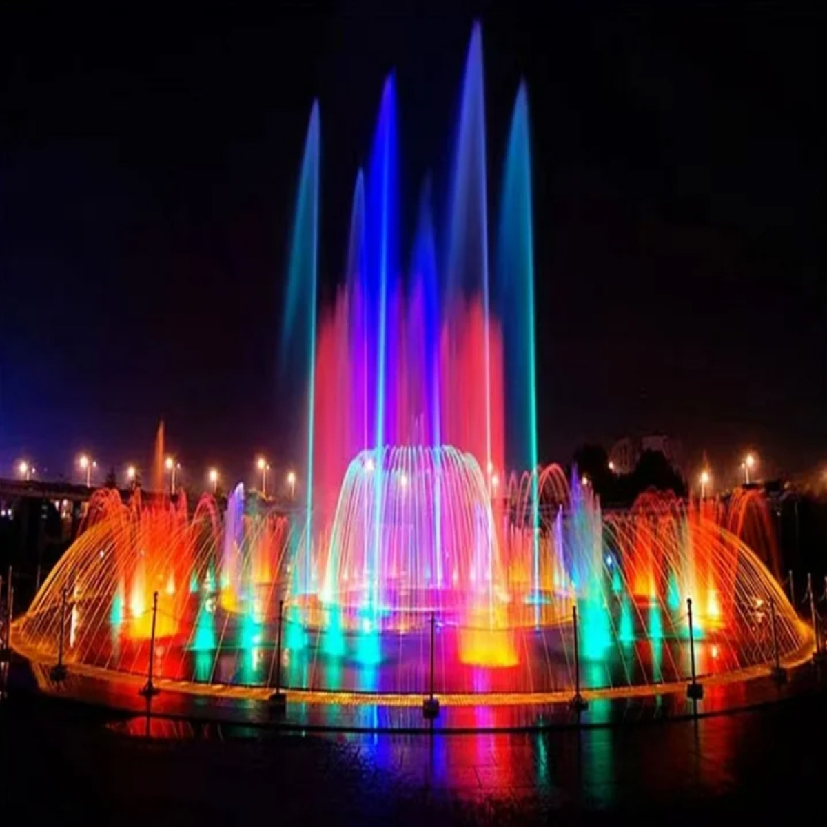 Ein farbenfroher Brunnen bei Nacht. Wasserstrahlen sind mit roten, orangen, grünen und blauen Lichtern beleuchtet, vor dunklem Himmel.