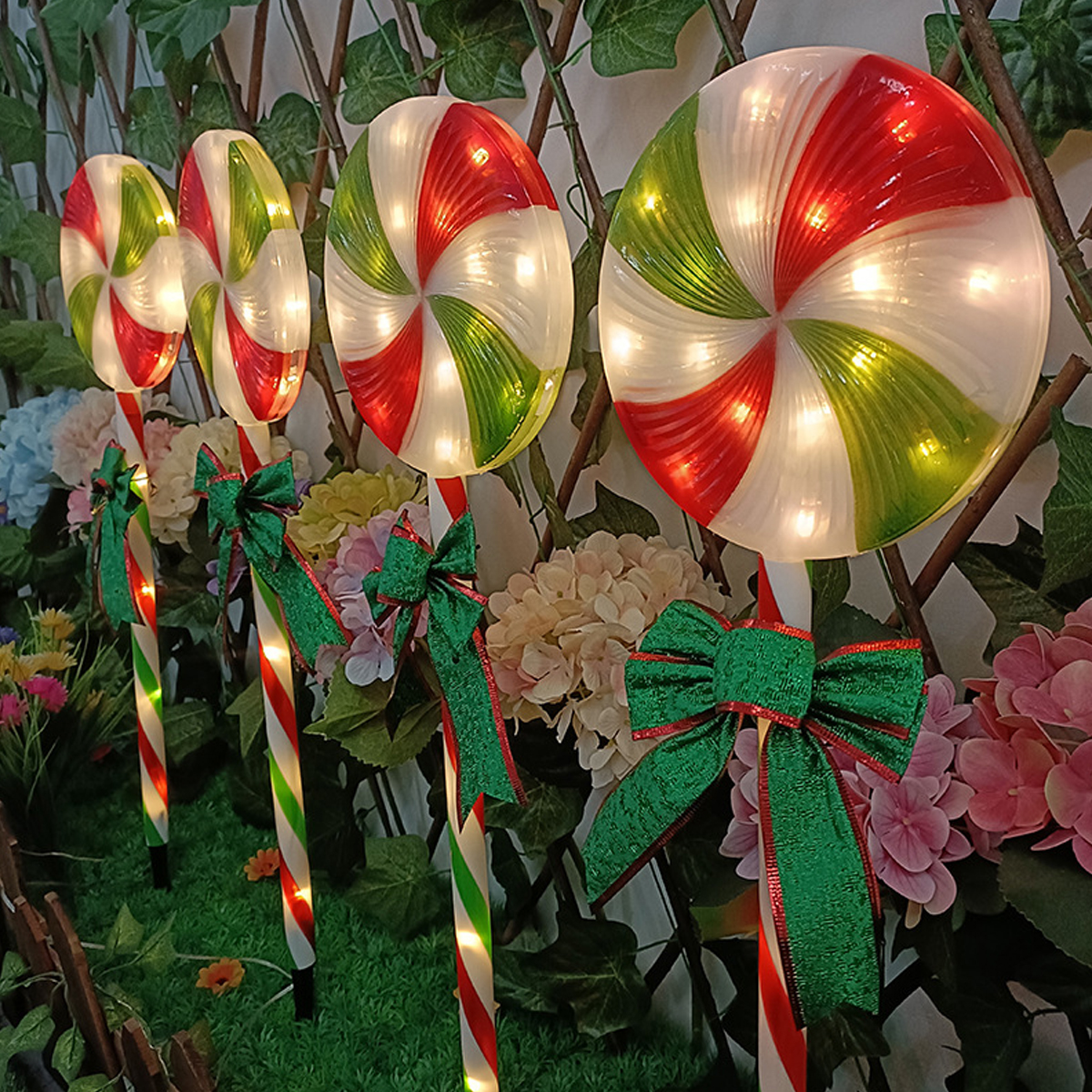 Vier beleuchtete Zuckerstangen-Dekorationen mit roten, grünen und weißen Wirbeln stehen in einem Garten, geschmückt mit Schleifen und Blumen.