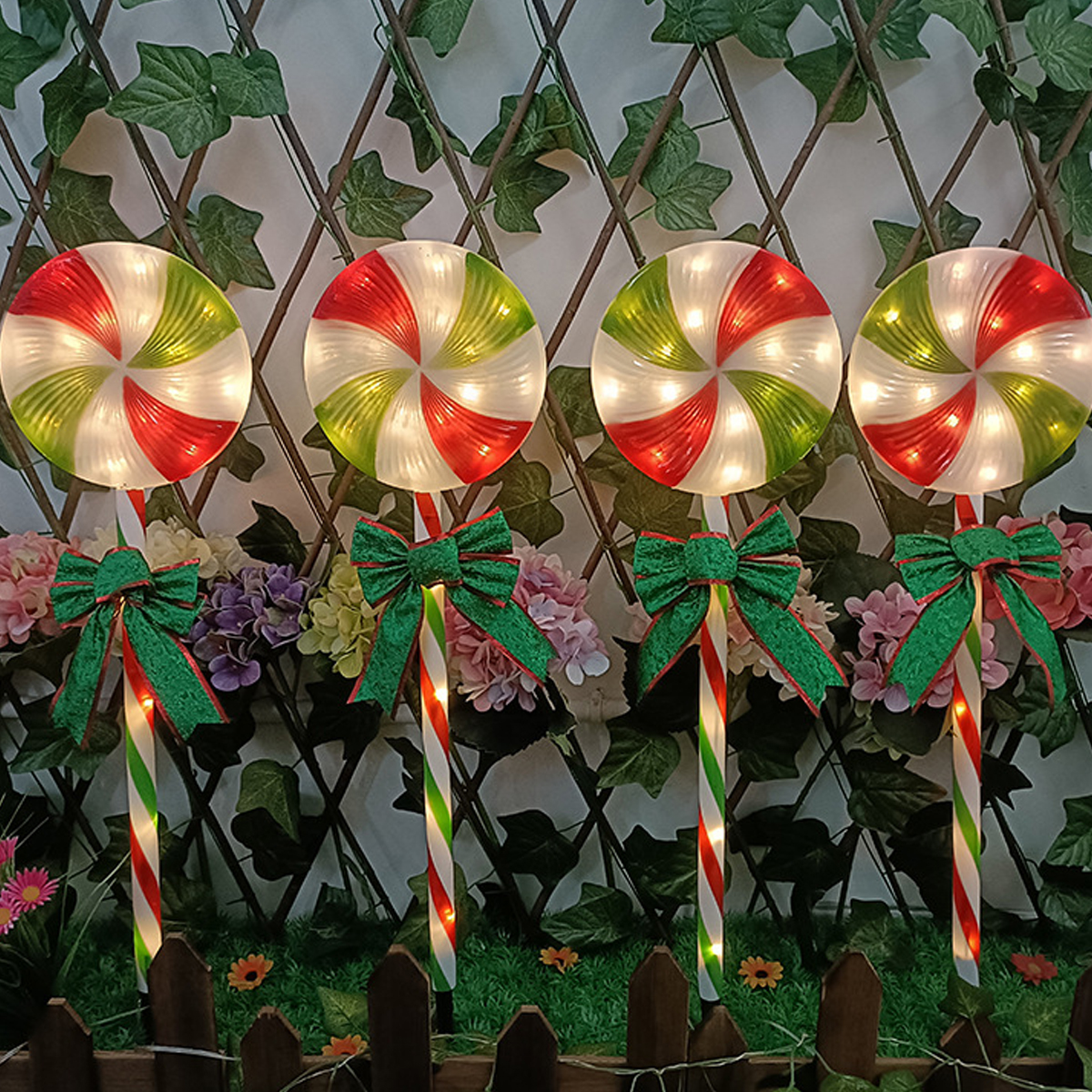 Vier beleuchtete Lutscher mit roten, grünen und weißen Spiralen. Sie haben grüne Schleifen und stehen vor Blumen und einem Zaun.
