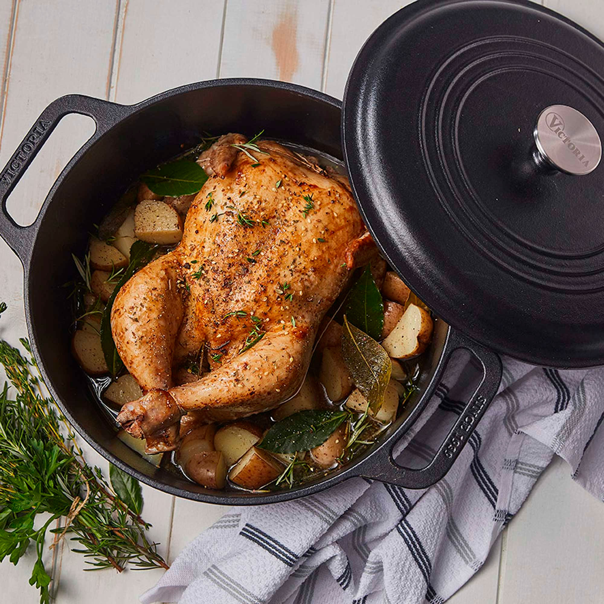 Pollo asado en olla negra con patatas, hierbas y tapa. Junto a una toalla blanca.