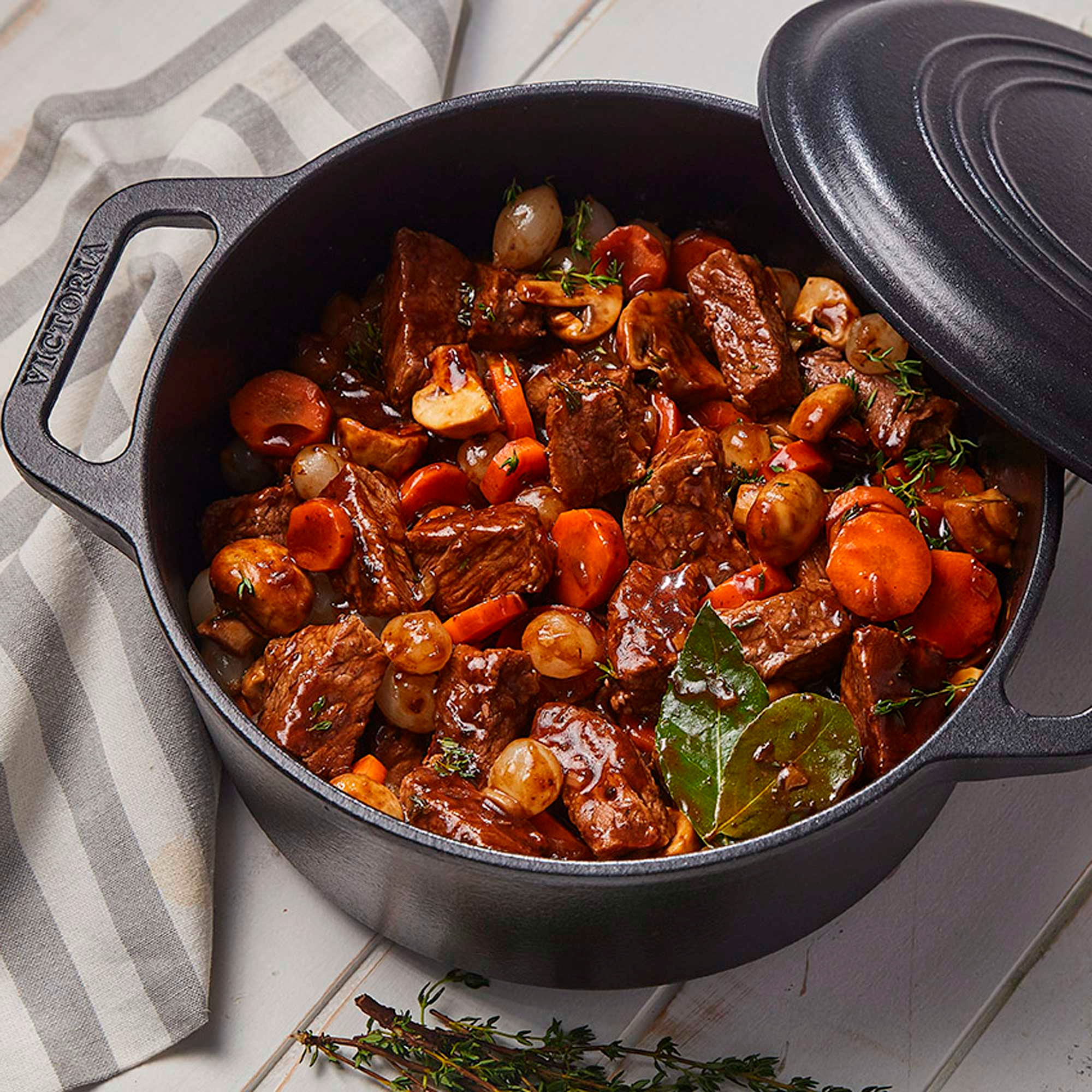 Estofado de ternera con zanahorias, champiñones y cebollas en una olla negra, tapa abierta. Servido sobre una mesa de madera blanca con una servilleta.